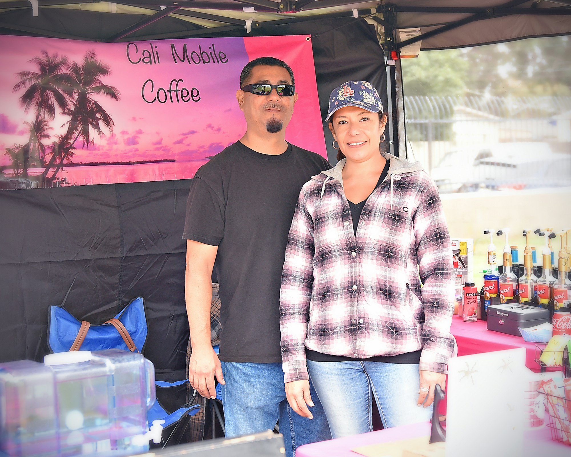 Two people at a mobile coffee stand with a "Cali Mobile Coffee" banner and syrup bottles on display.