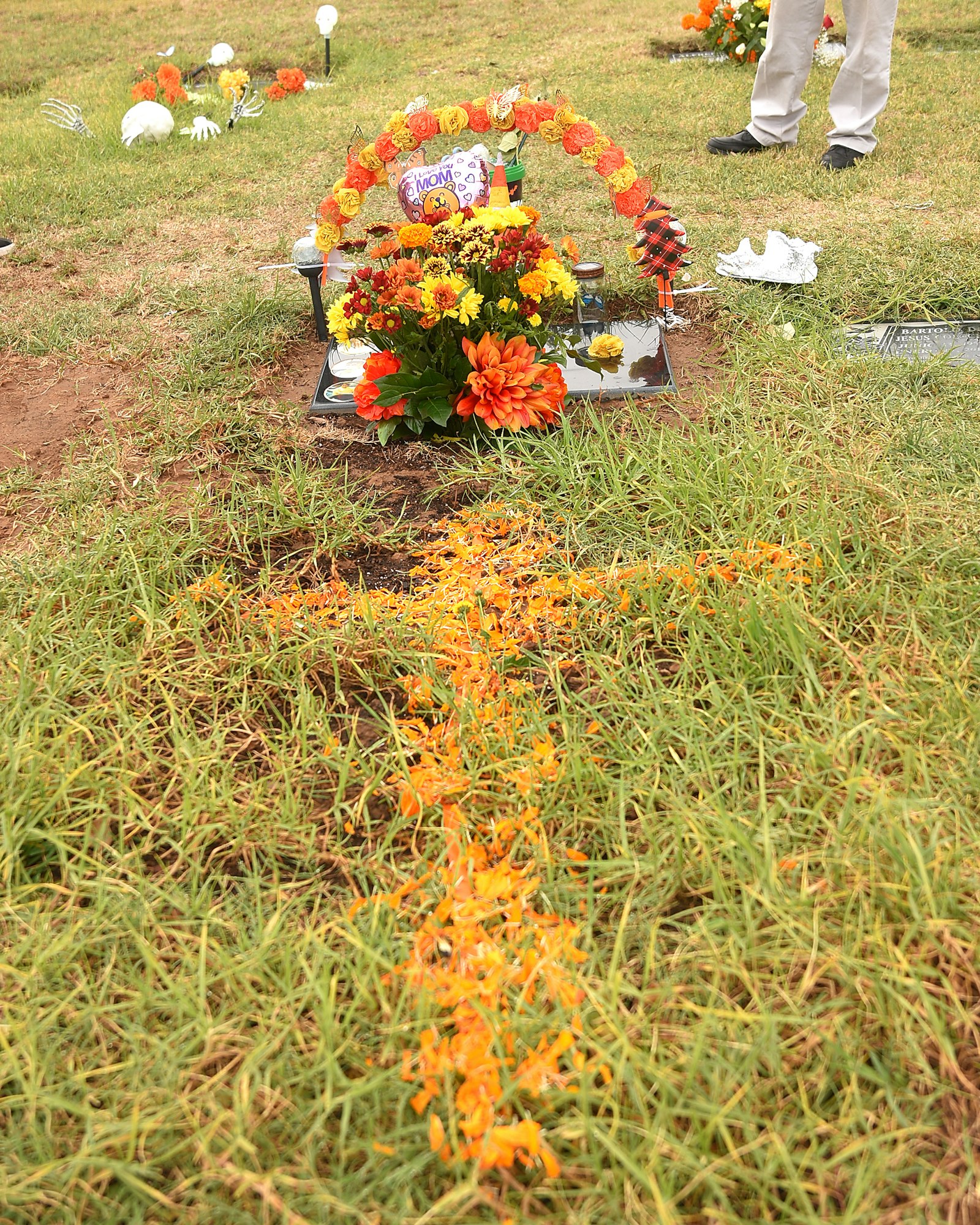 A gravesite with floral decorations, a balloon, and a trail of orange petals, with a person's legs visible in the background.