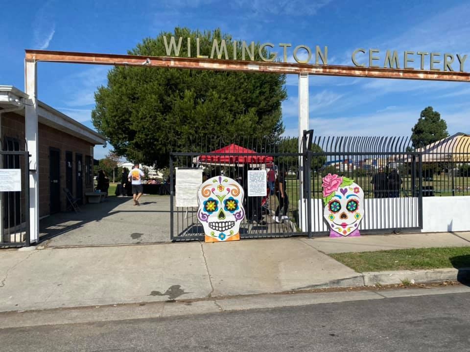 The image shows the entrance to Wilmington Cemetery, decorated with colorful skull artwork and a festive atmosphere.