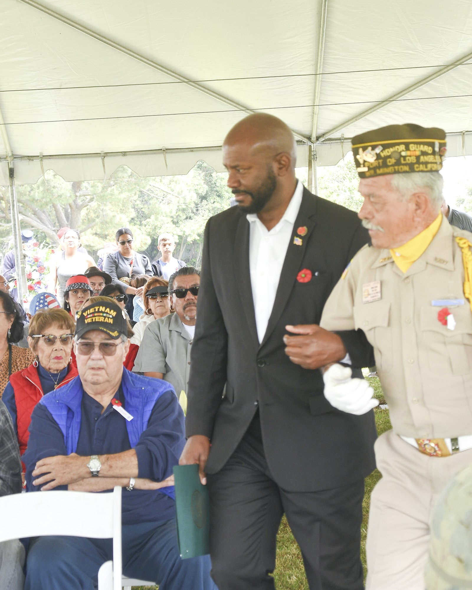 Two men in ceremonial attire walking, audience seated under tent, outdoor event, military or veterans theme.