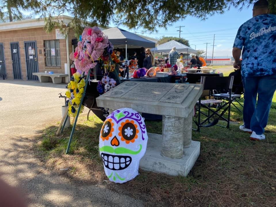 The image features a decorated area with a colorful sugar skull, flowers, and people gathered around. It suggests a festive occasion.