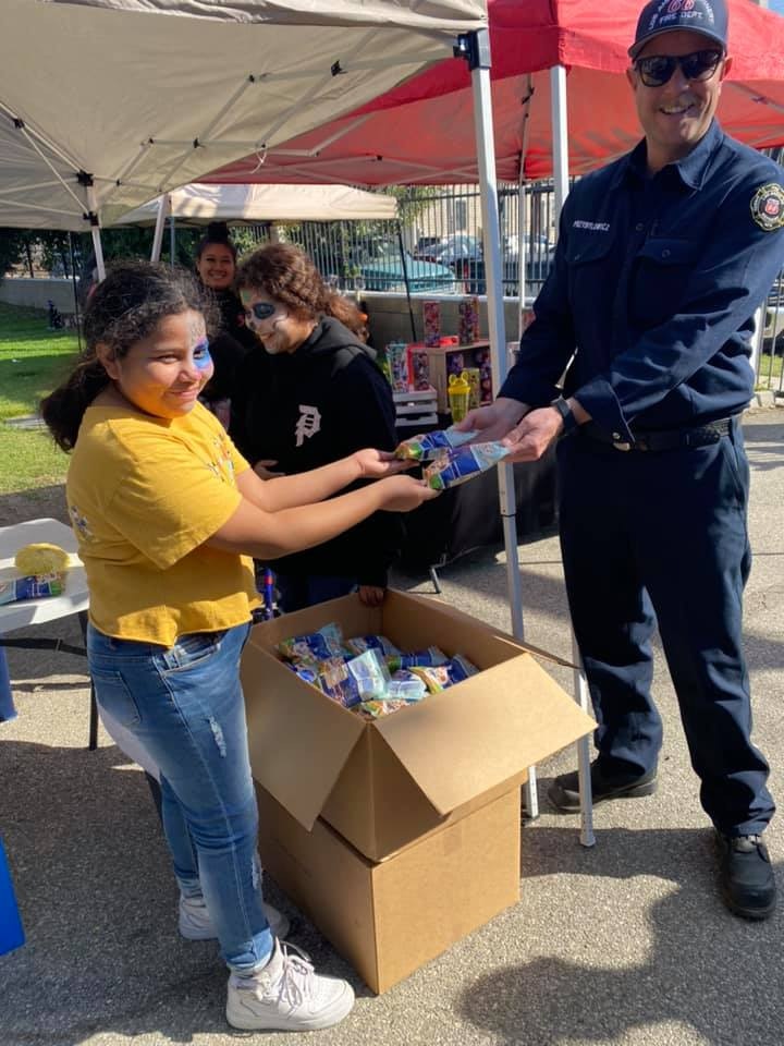 A child receives a small item from a firefighter at an outdoor event with tents, showing community engagement and support.
