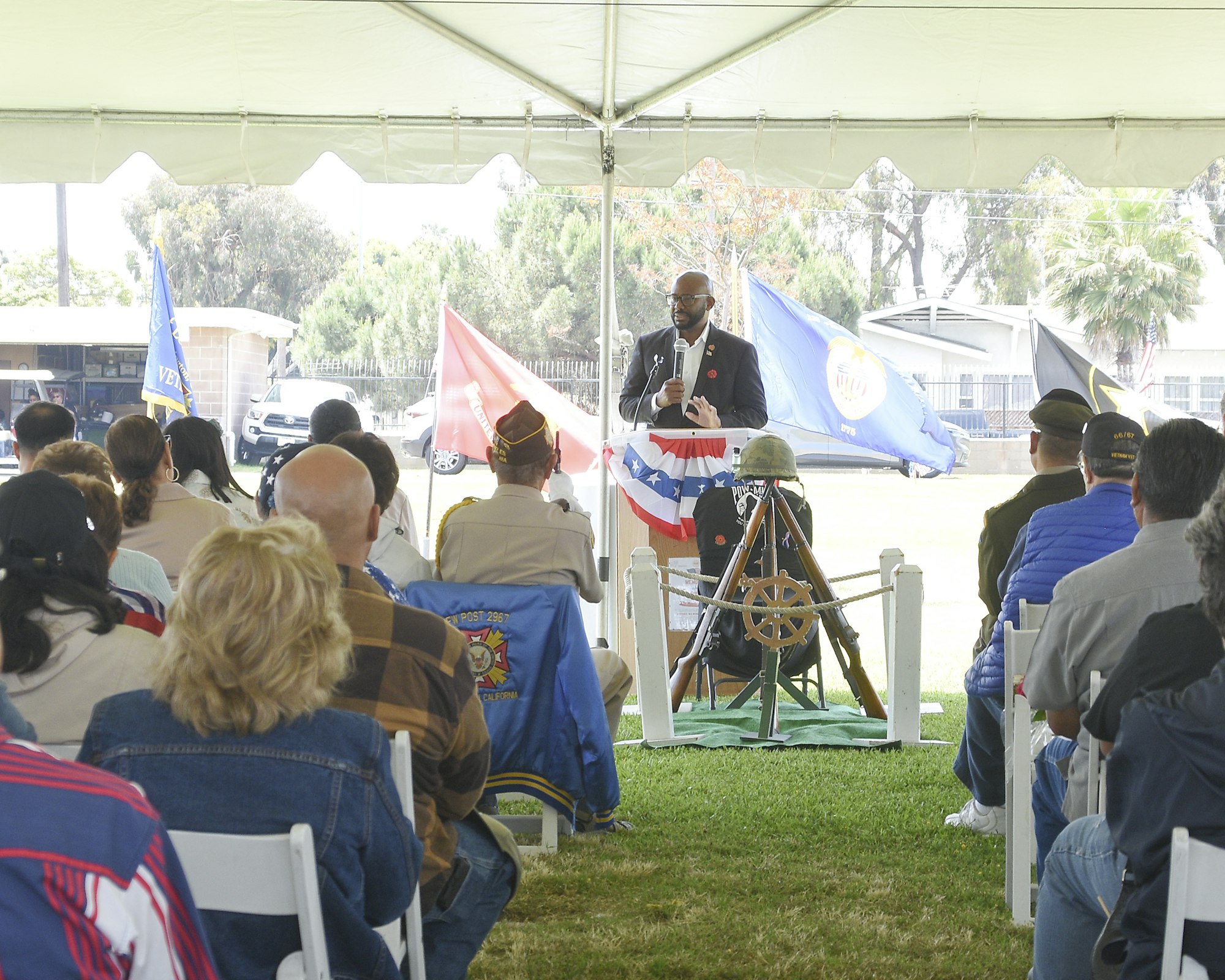 A speaker at an outdoor event with an audience under a tent, military uniforms, flags, and a ship's wheel on display.