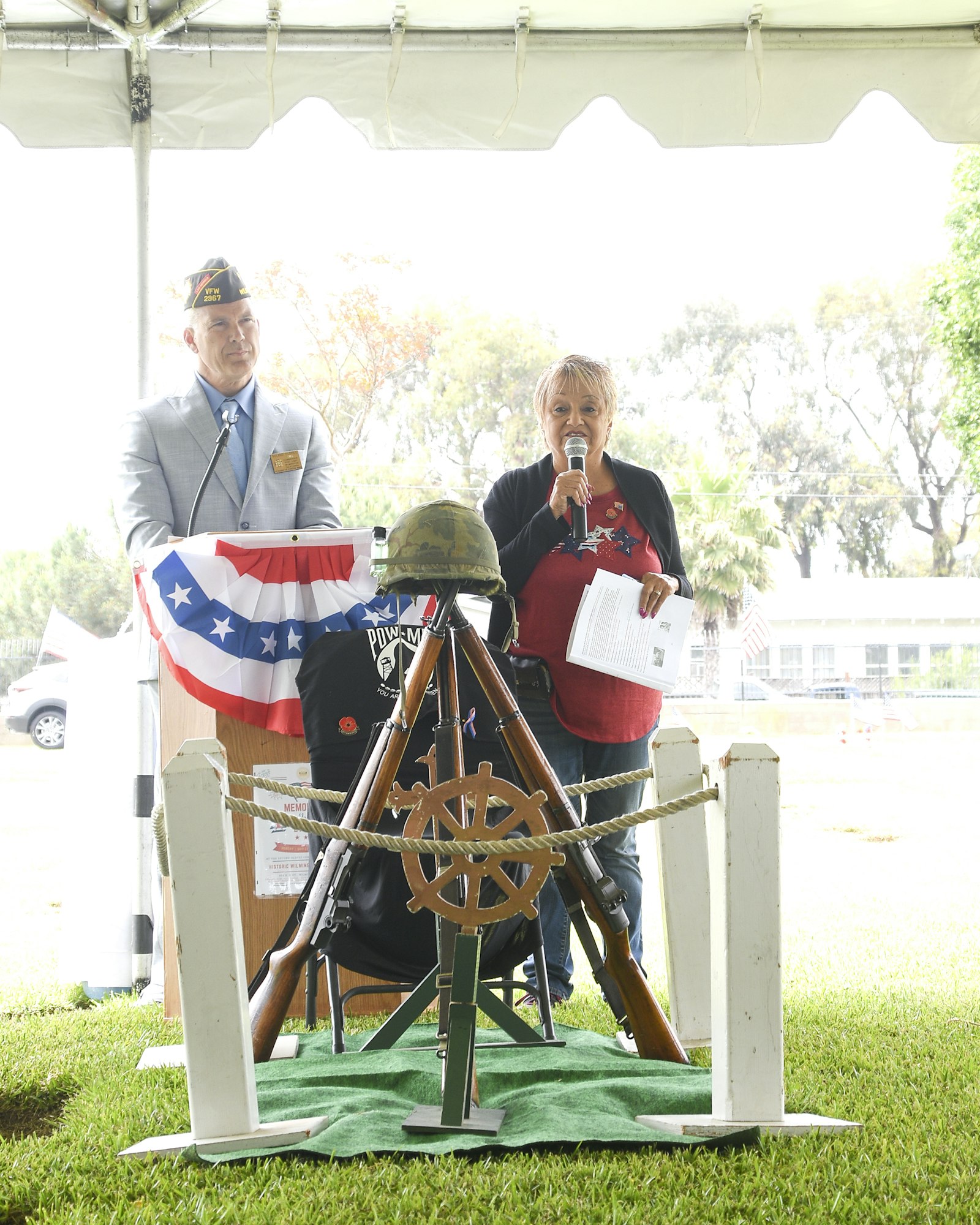 A ceremony with two people, one speaking, military symbols, a POW/MIA flag, and a ship's helm in the foreground.