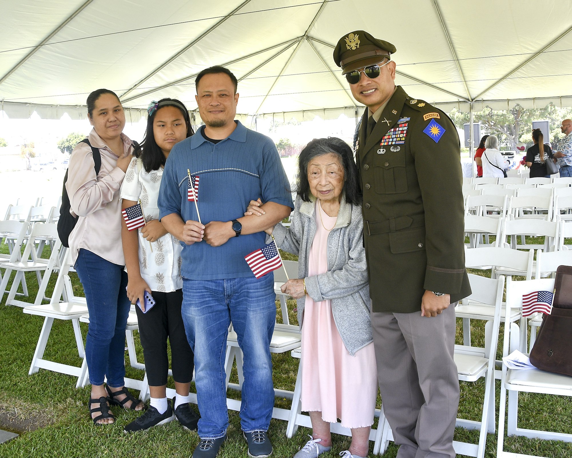 A family posing with a military officer at an event, holding American flags, with rows of chairs in the background.