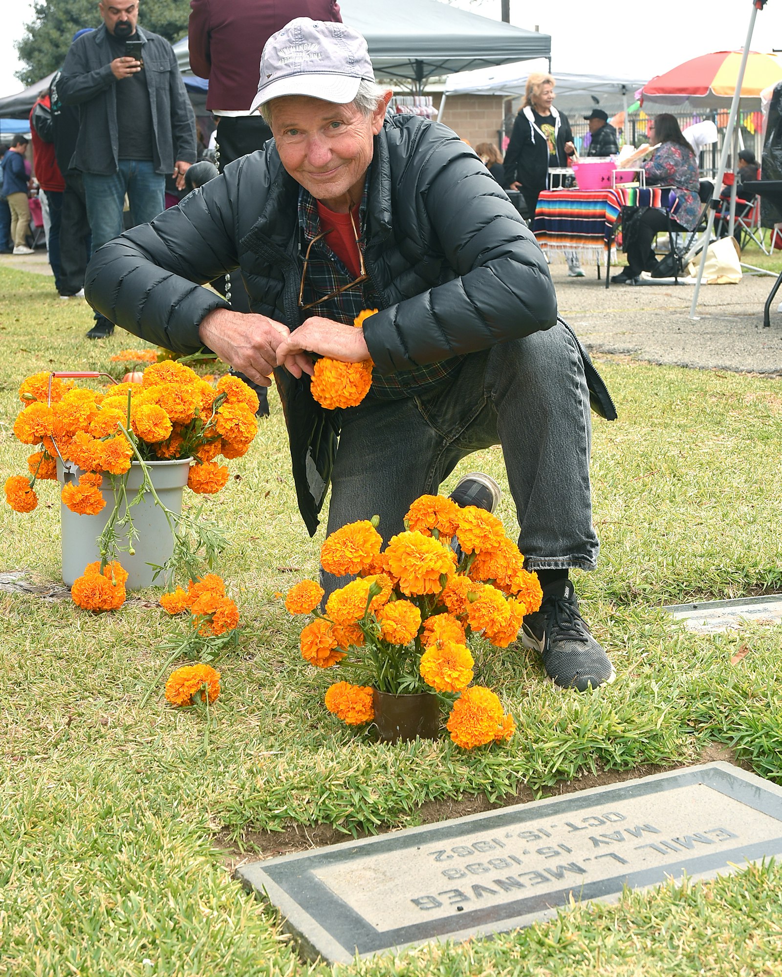 A person crouching by vibrant marigolds at a gravesite, with people and tents in the background.