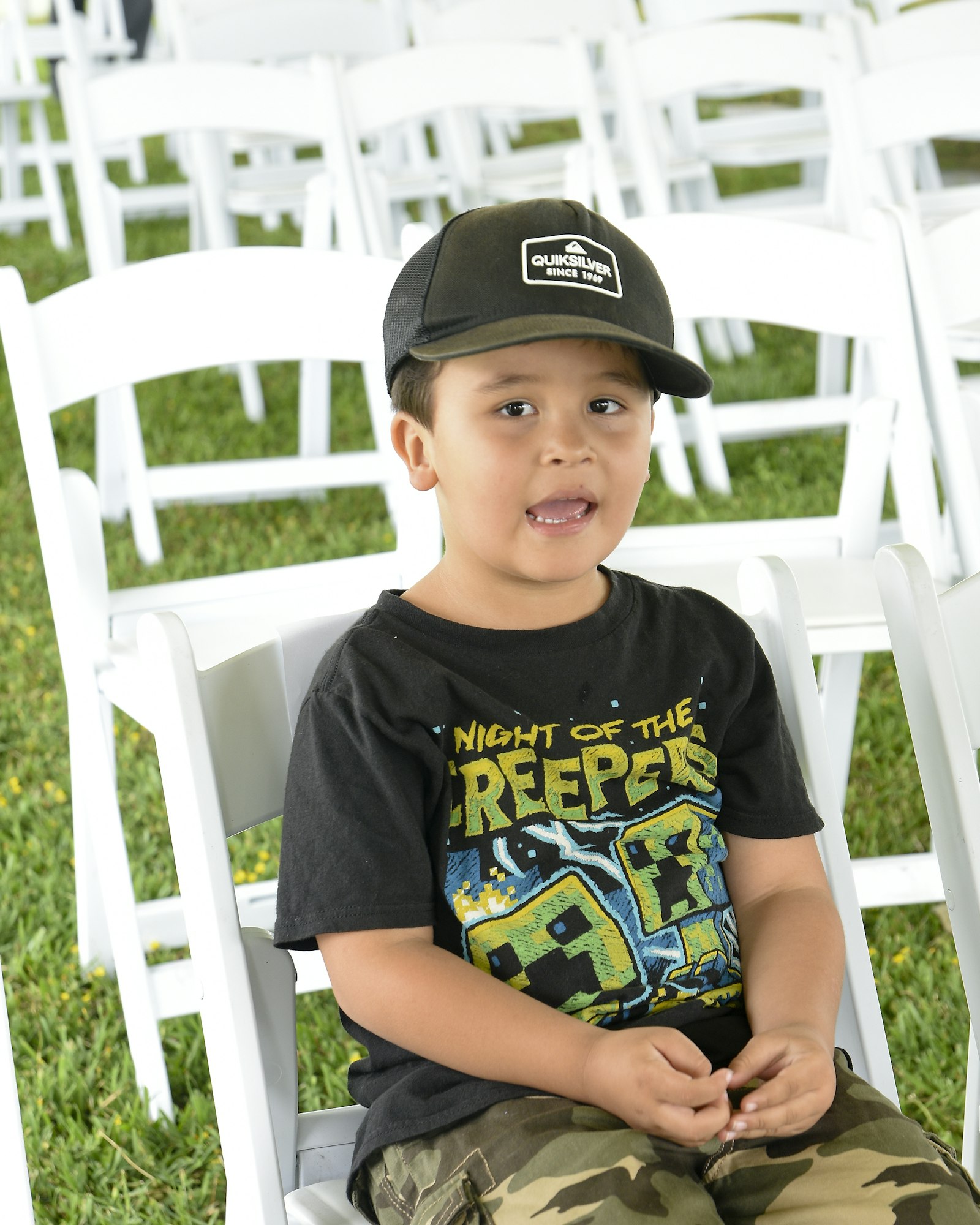 May contain: baseball cap, cap, clothing, hat, t-shirt, grass, plant, boy, child, male, person, chair, furniture, face, head, photography, portrait, and people