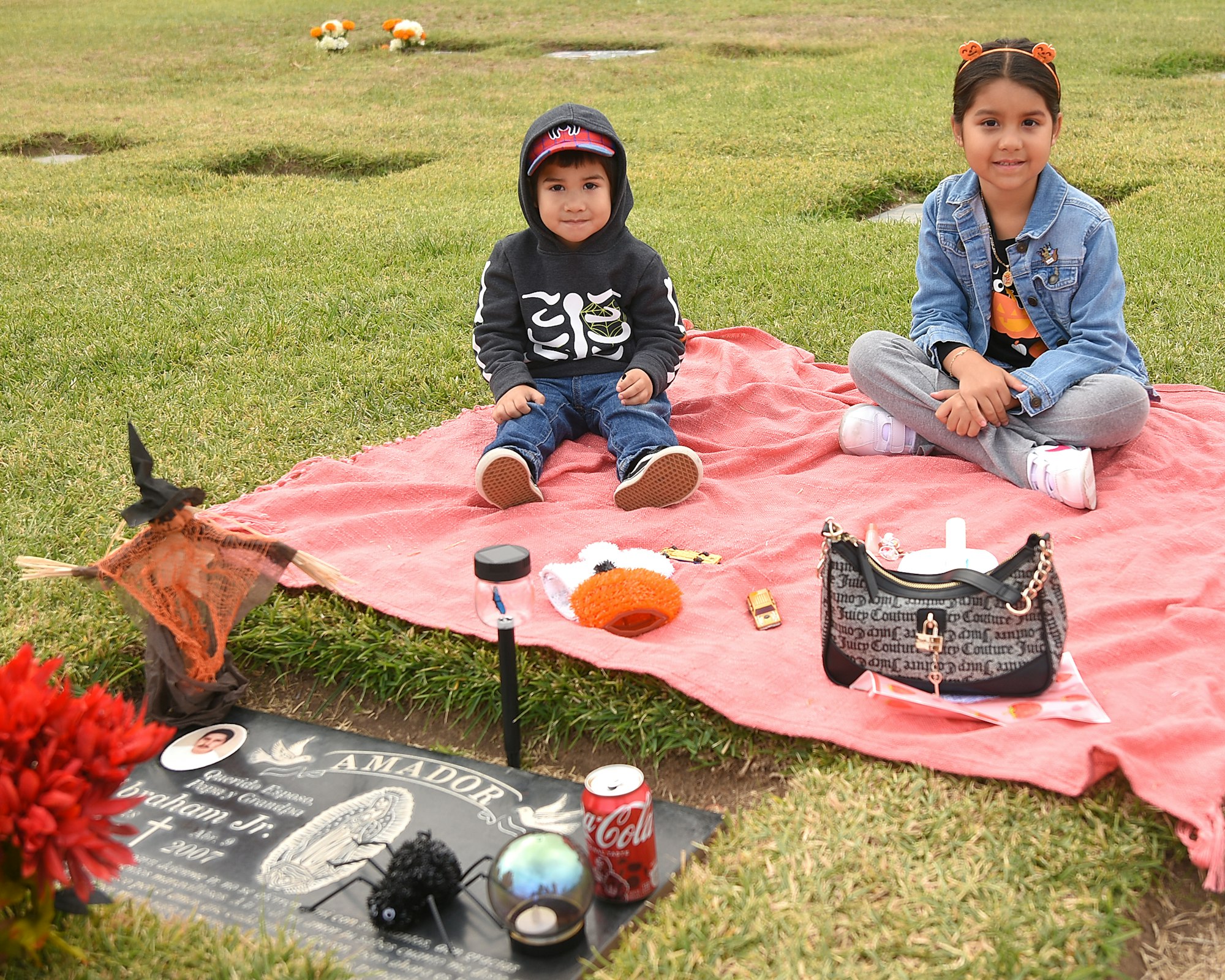 Two children sitting on a blanket by a decorated gravesite with flowers, beverages, and personal items.