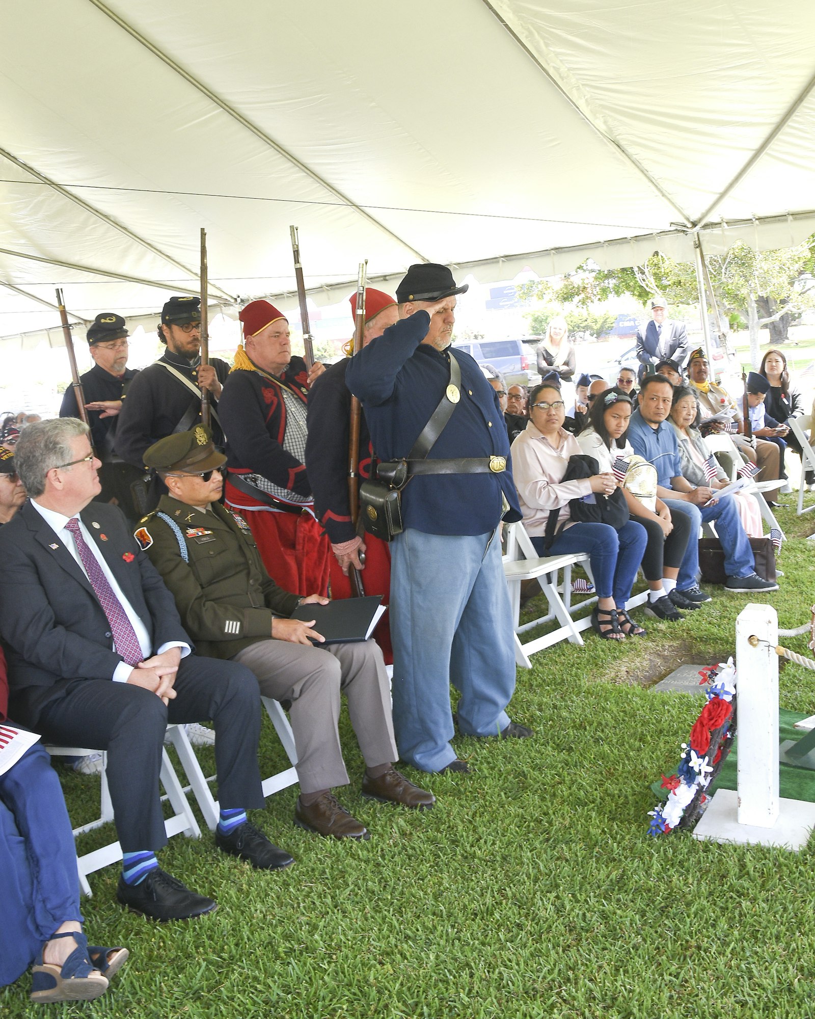 A group of people in historical military uniforms standing near seated attendees under a tent.