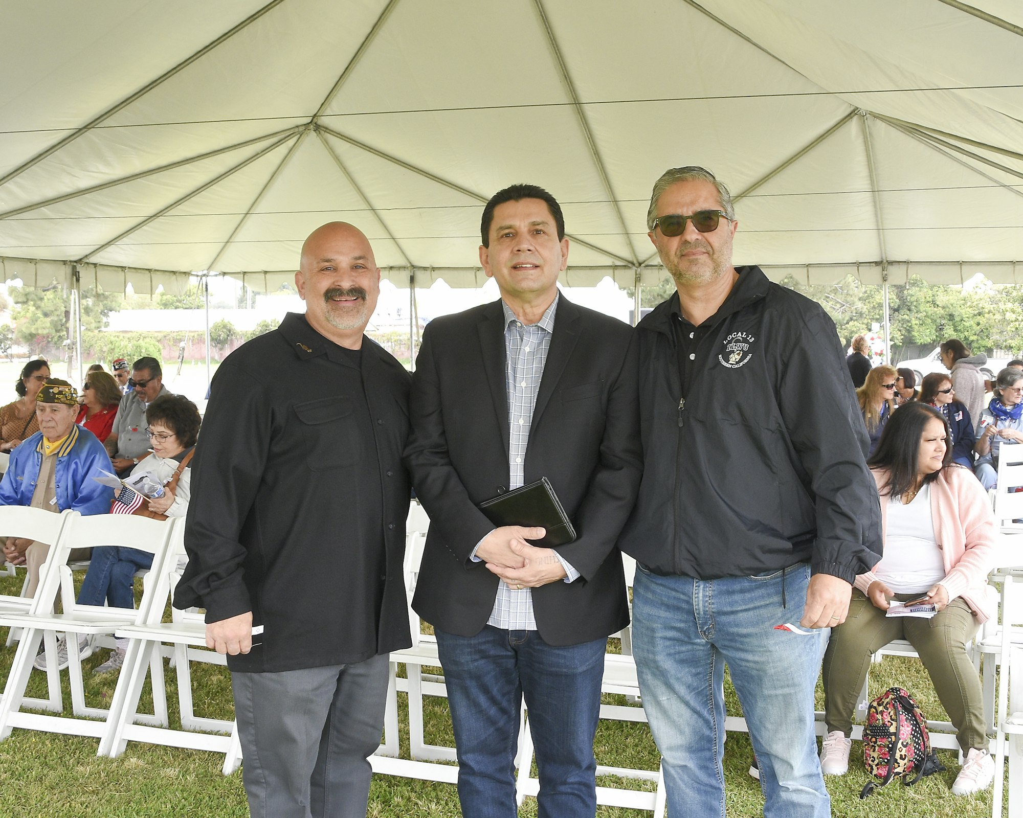 Three men posing for a photo under a tent, with seated people in the background, possibly at an outdoor event.