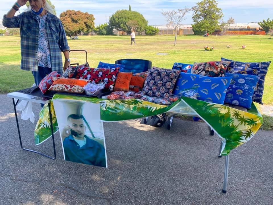 A person stands by a table displaying various colorful pillows and a photo, set up in a park.