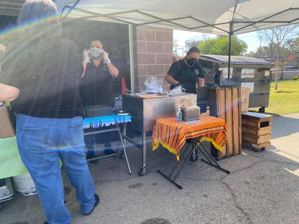 Two people are preparing food at a food stall outside, wearing masks, with tables and cooking equipment visible.
