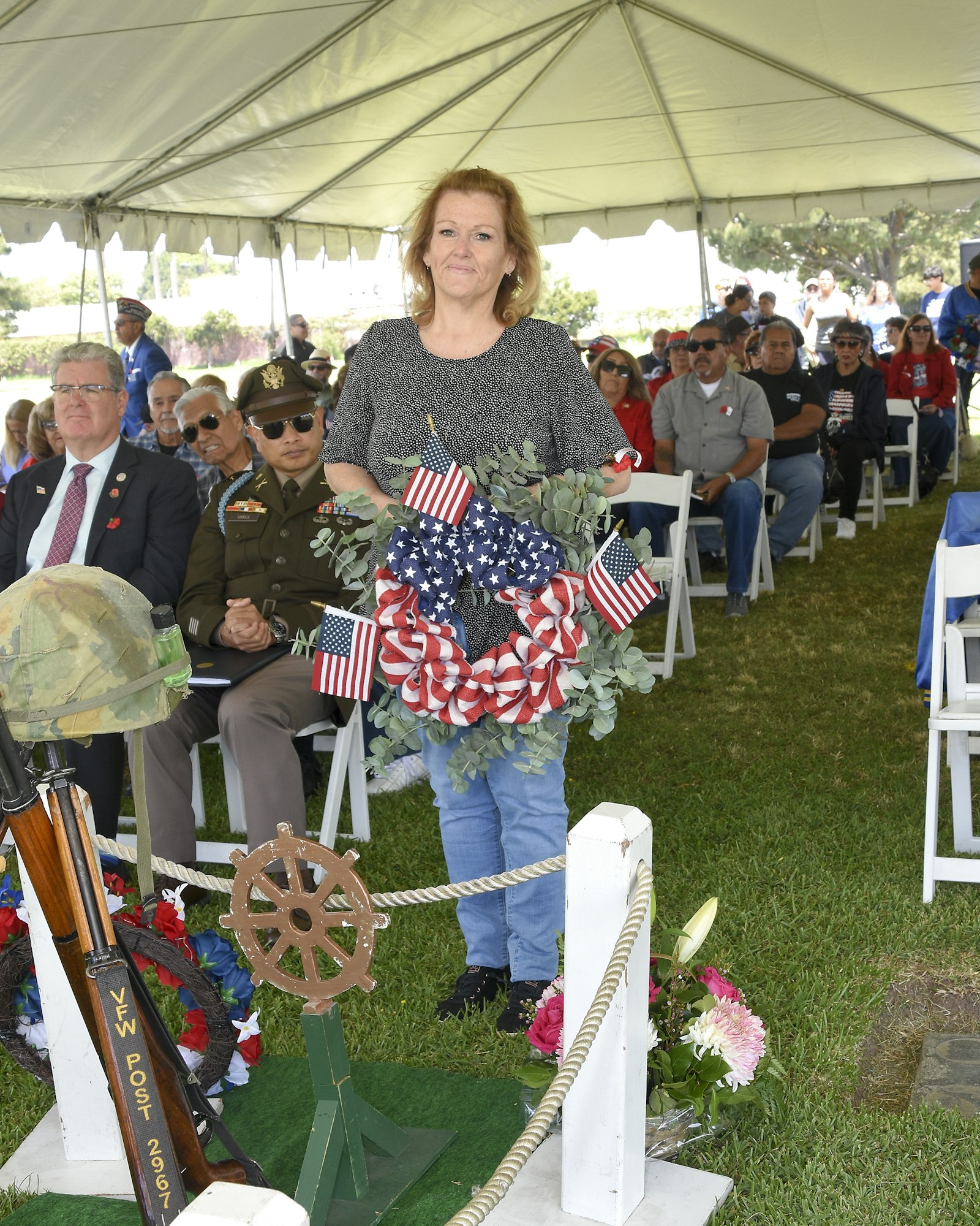 A woman standing behind a memorial wreath with flags at an outdoor commemorative event with onlookers.