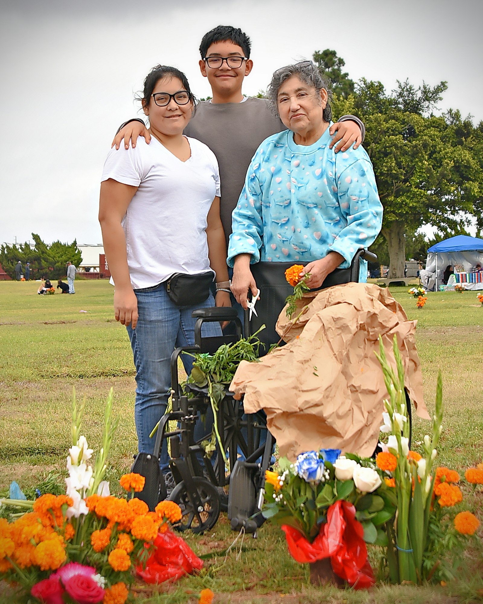 A family posing together in a park with flowers and a wheelchair, hinting at a memorial or a visit to a cemetery.