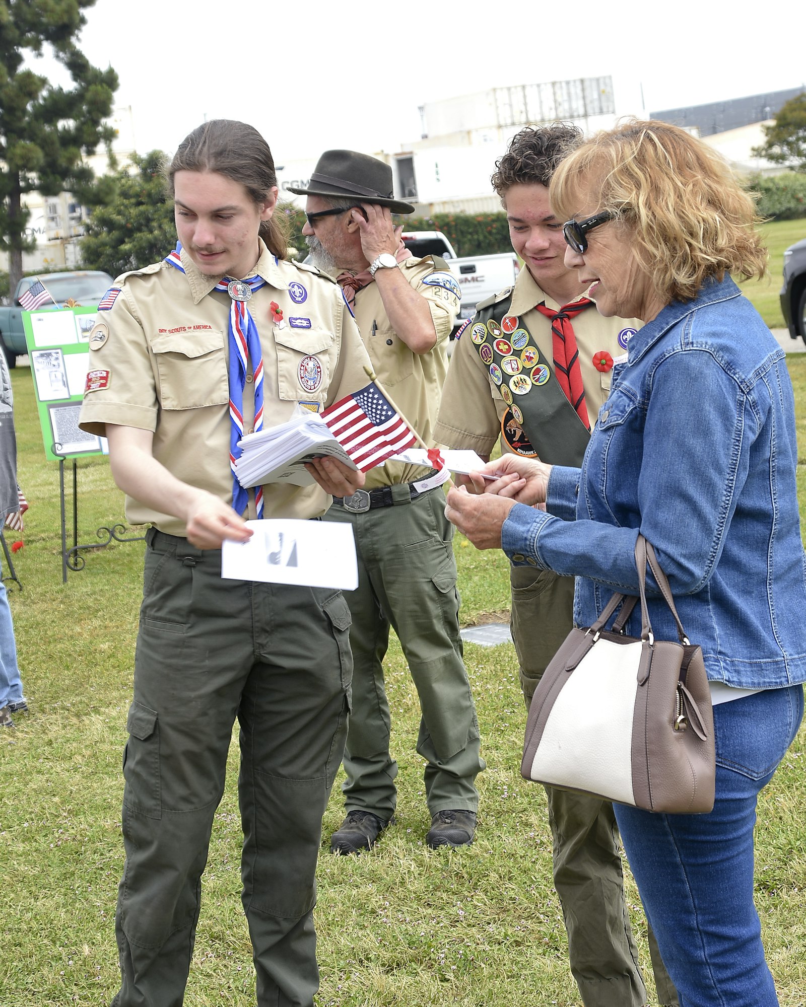 Scouts in uniform with badges, one holding flags, interacting with adults outdoors.