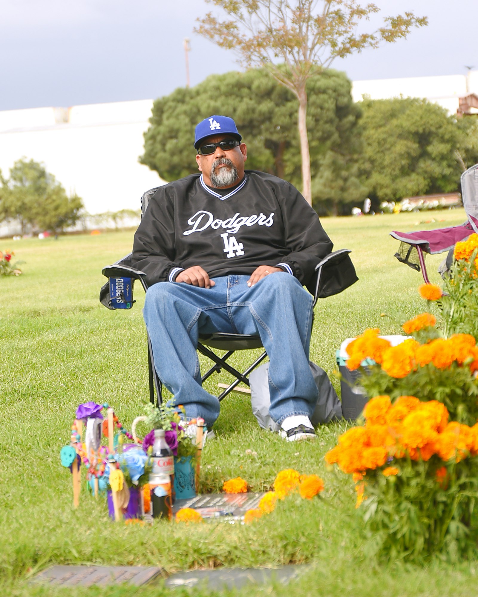 Man in Dodgers jacket sitting by floral offerings in a park-like setting.