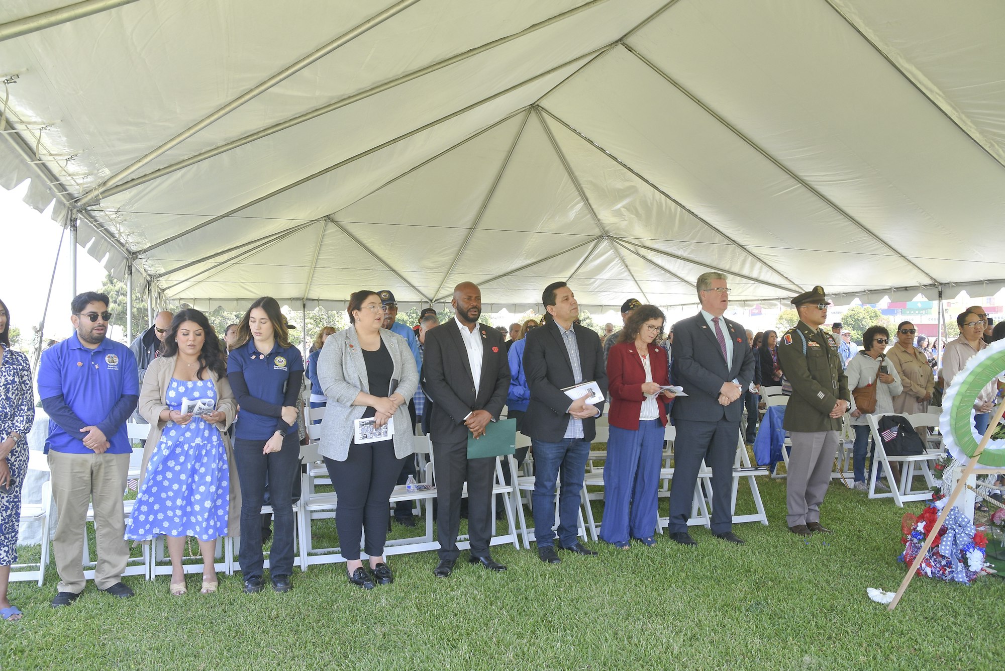A group of people standing solemnly under a tent, likely at an outdoor event or ceremony.