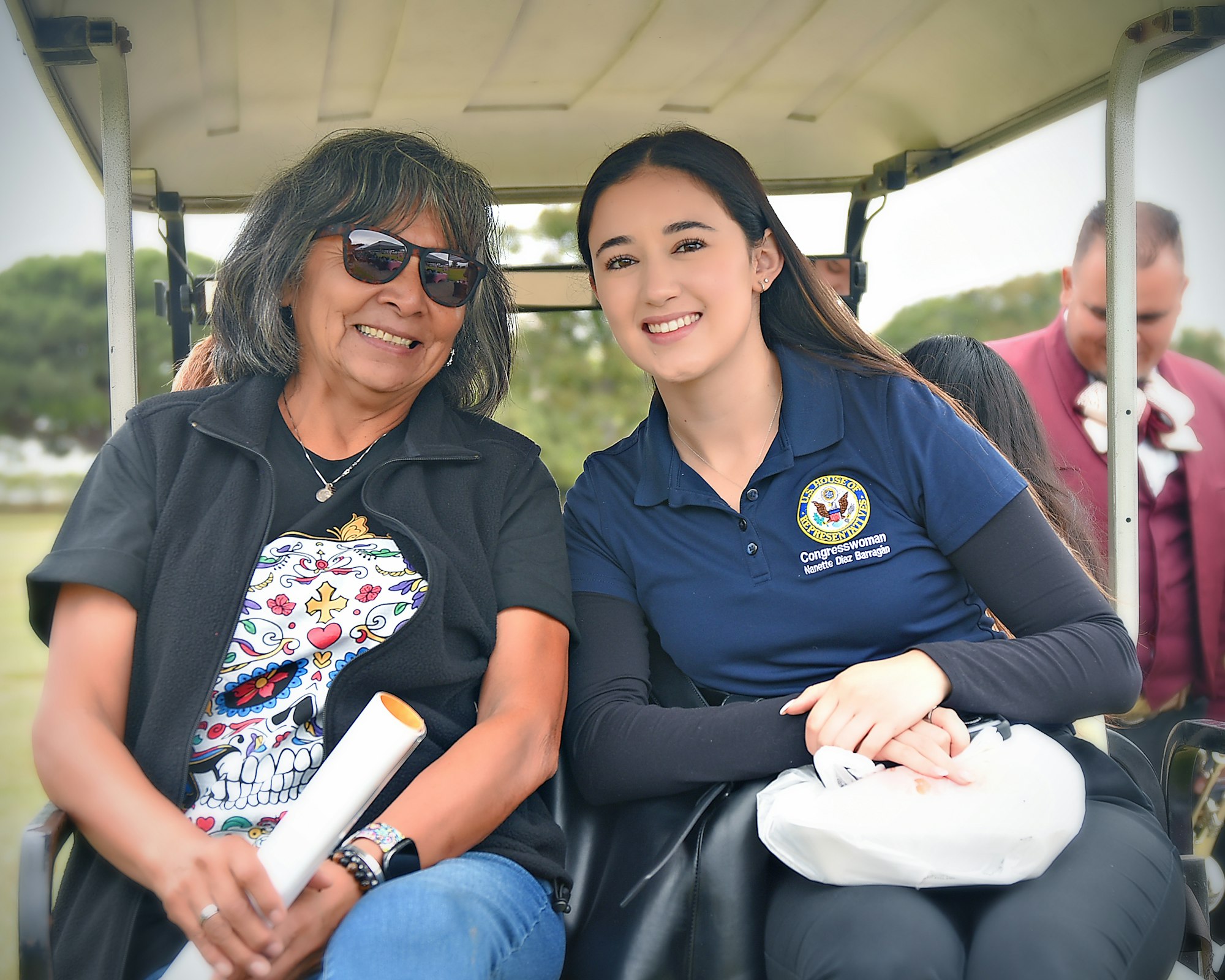 Two women smiling on a golf cart, one with a "Congresswoman" shirt. There's a sunny outdoor setting.