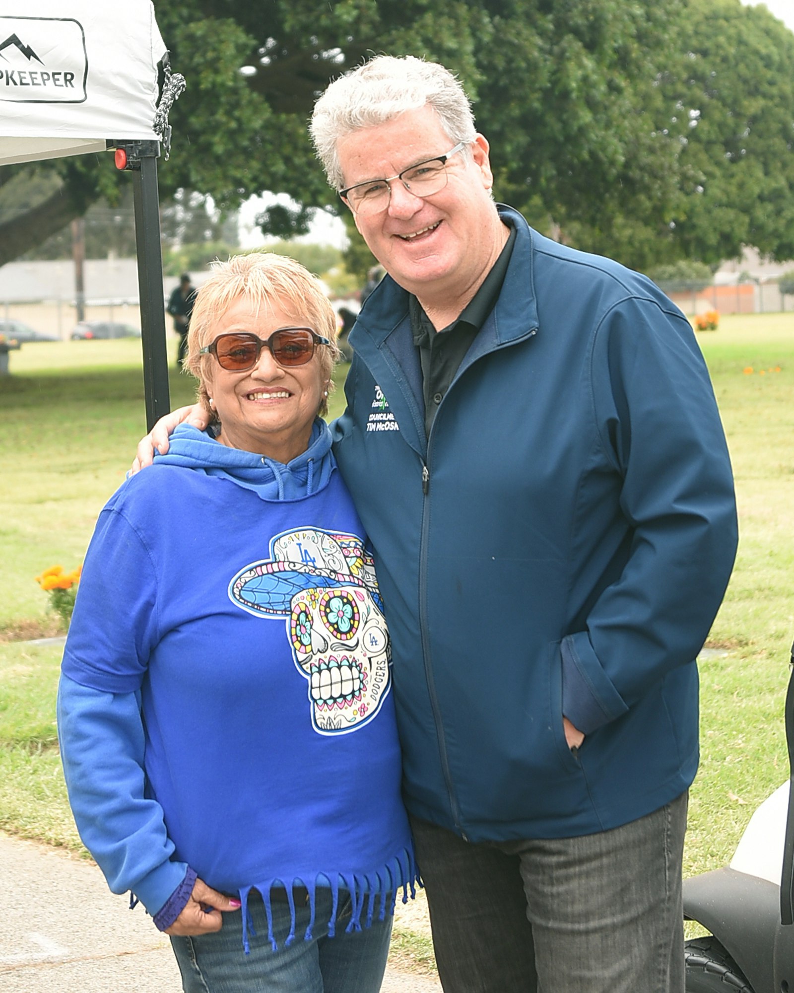 A man and a woman smiling, standing next to each other outdoors, woman in blue hoodie with design, man in blue jacket.