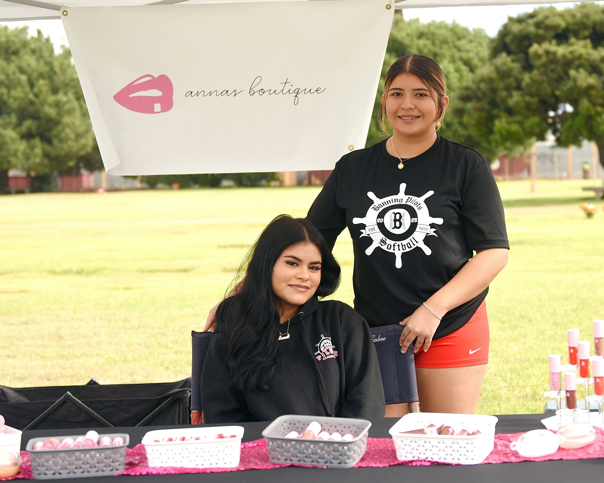 Two individuals at a boutique stand with beauty products on a table, under a banner.