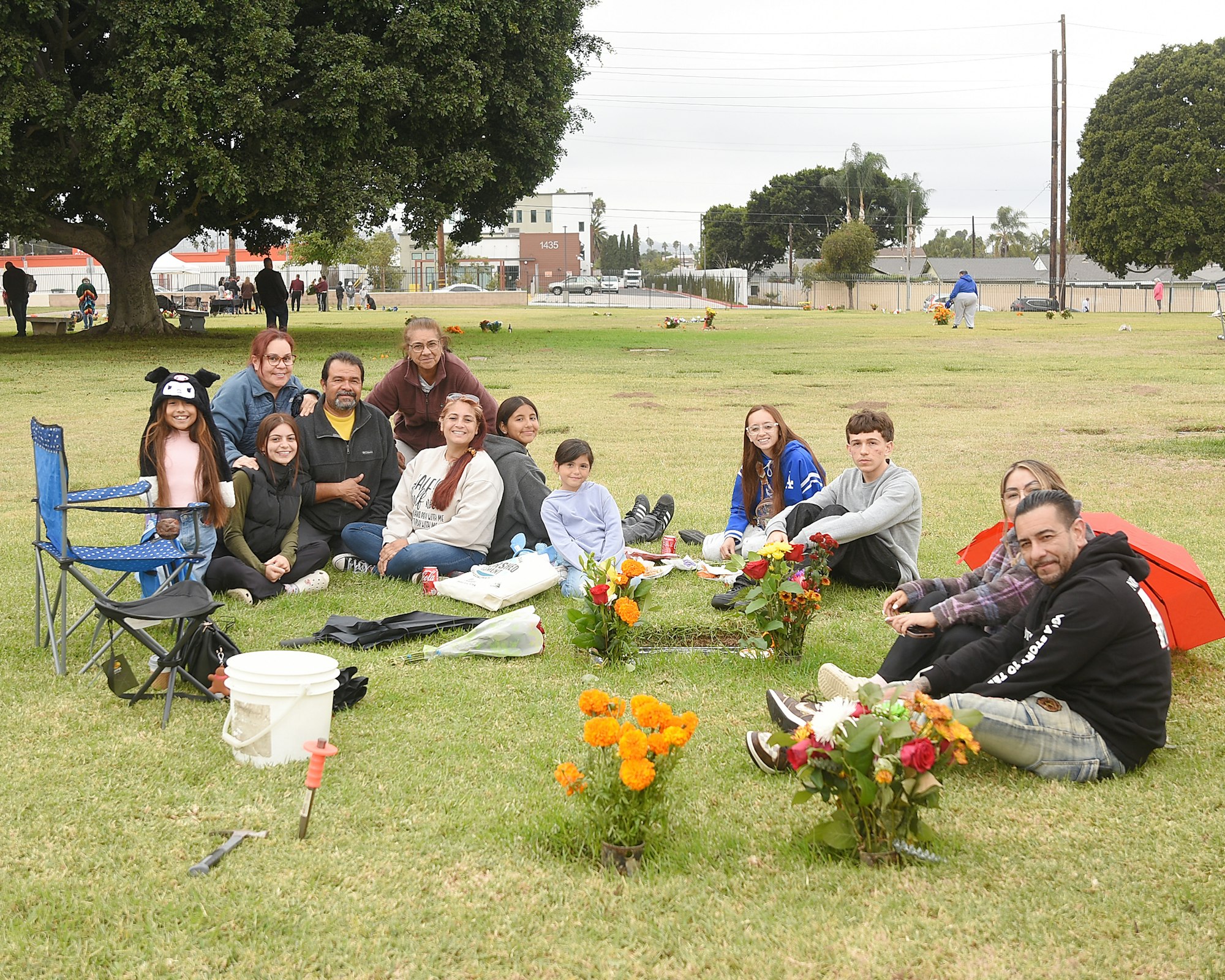 Group of people sitting on grass with flowers, possibly visiting a gravesite or commemorating an event, casual, outdoors.