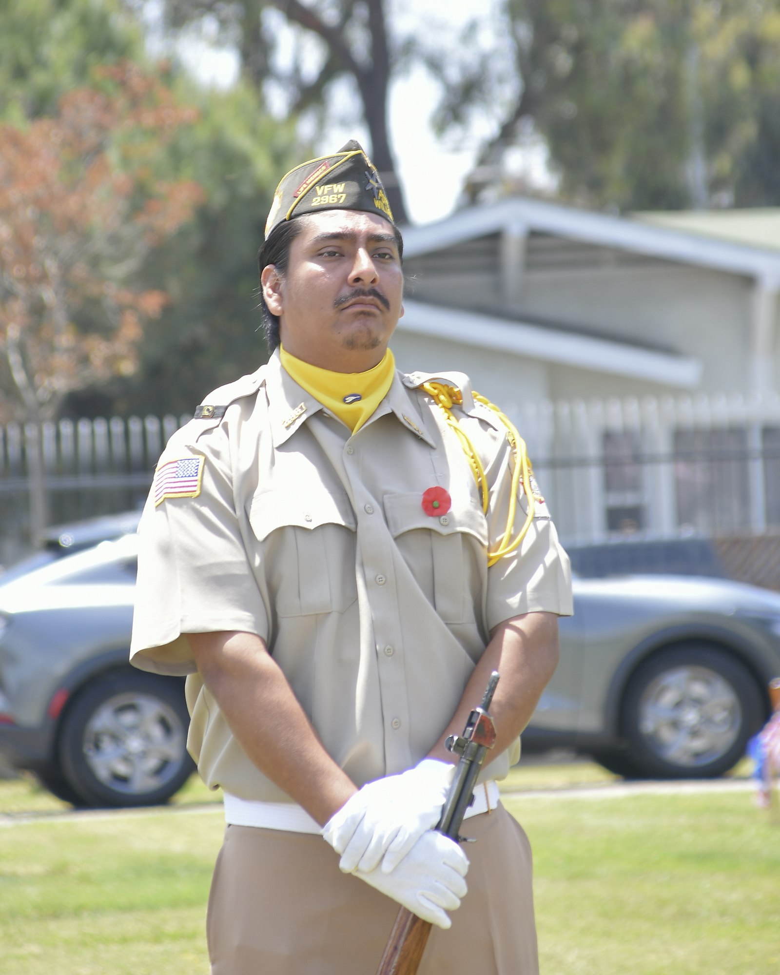 A person in uniform with a cap and gloves, holding a rifle, with a VFW insignia on the cap.