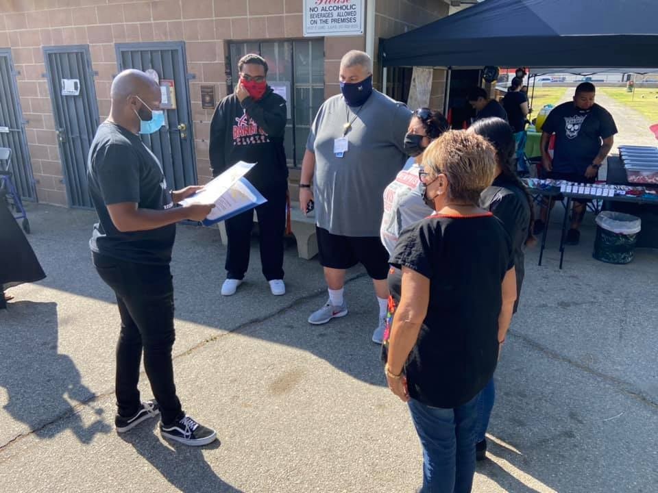 A group of people gathered outdoors, some wearing masks, discussing something while a person holds a document.