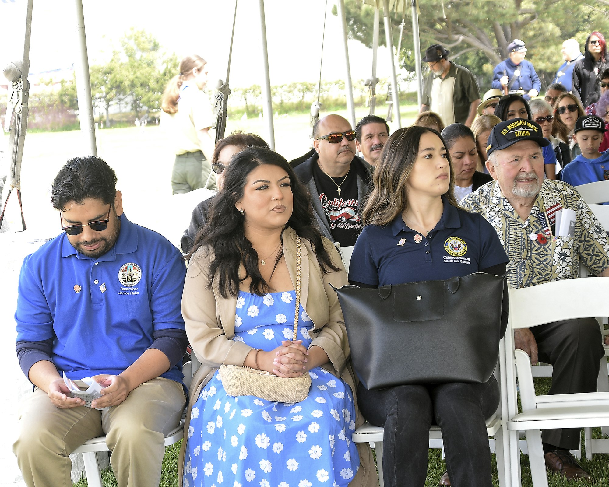 People sitting in chairs at an outdoor event, some wearing badges or emblems, indicating a formal or ceremonial gathering.