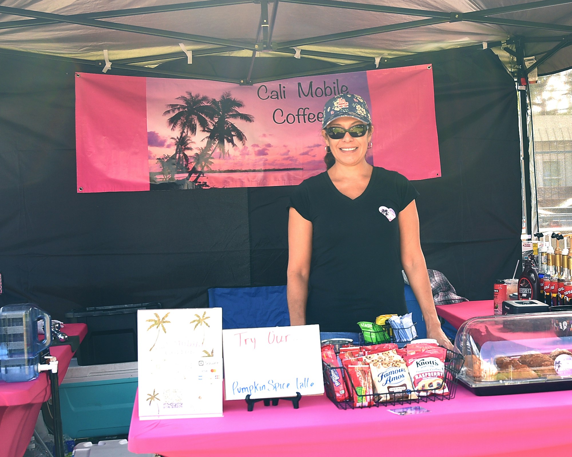 A smiling person stands at a booth with a "Cali Mobile Coffee" banner, snacks, coffee menu, and a pink tablecloth.