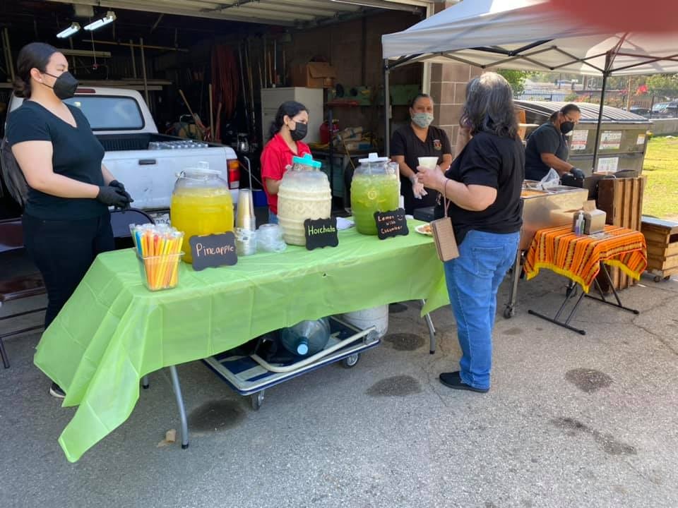 A outdoor drink stand selling beverages like pineapple, horchata, and lemonade, with people interacting and serving.