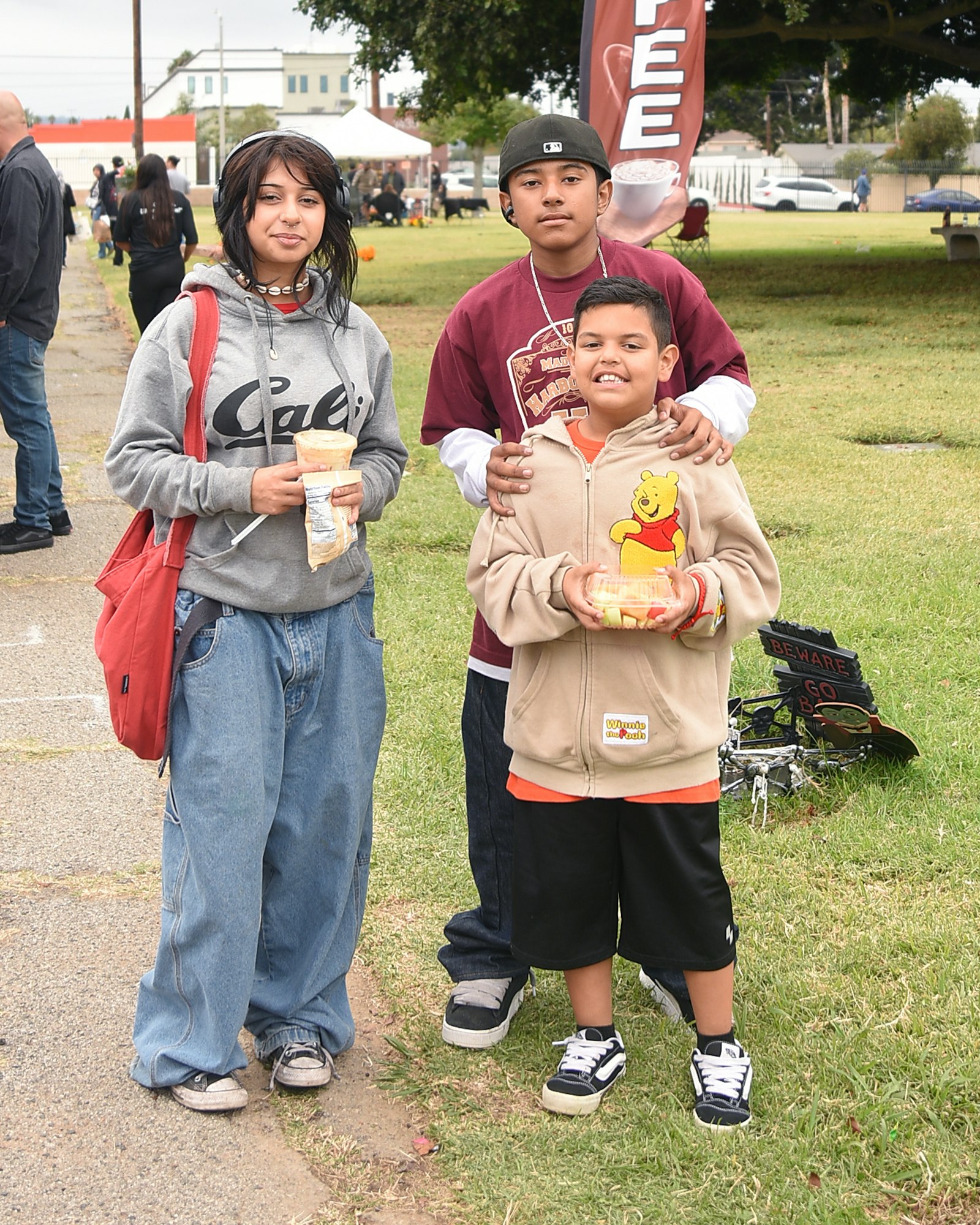 Three people in casual attire holding snacks, standing on grass with a festival background.