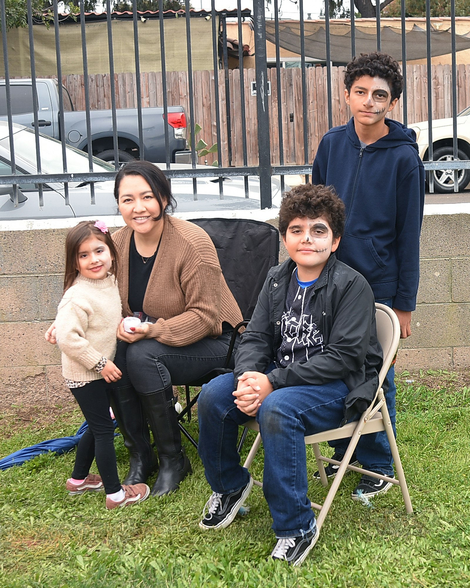 A group of people sitting outside, two children with face paint, and cars in the background.