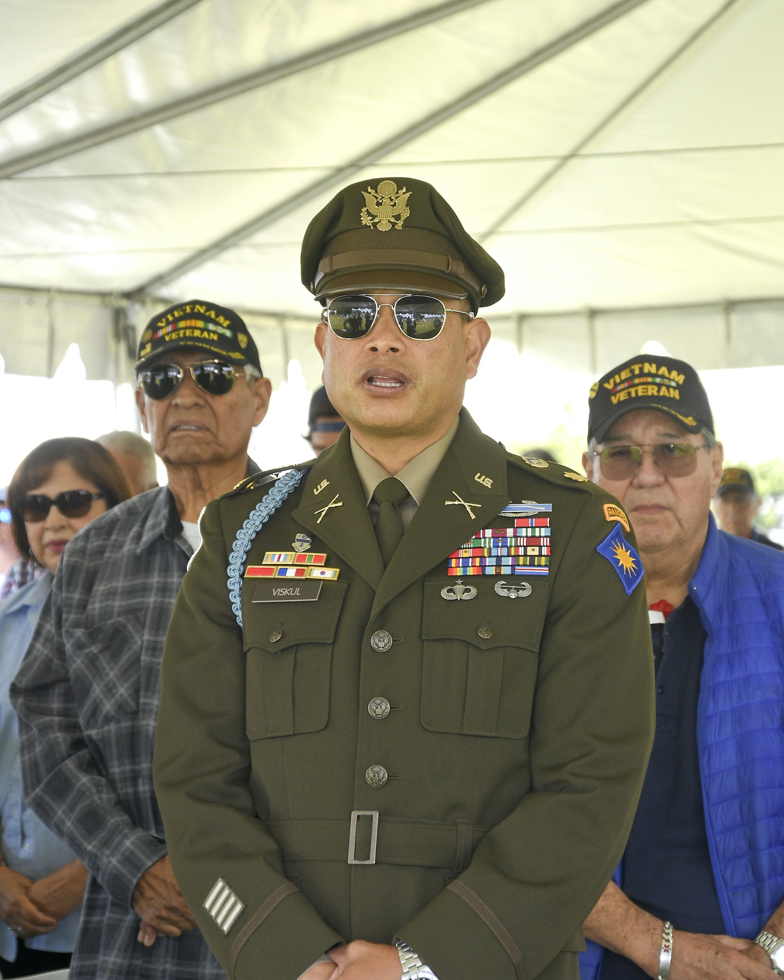A uniformed military officer with medals stands in front, with two older veterans in the background.