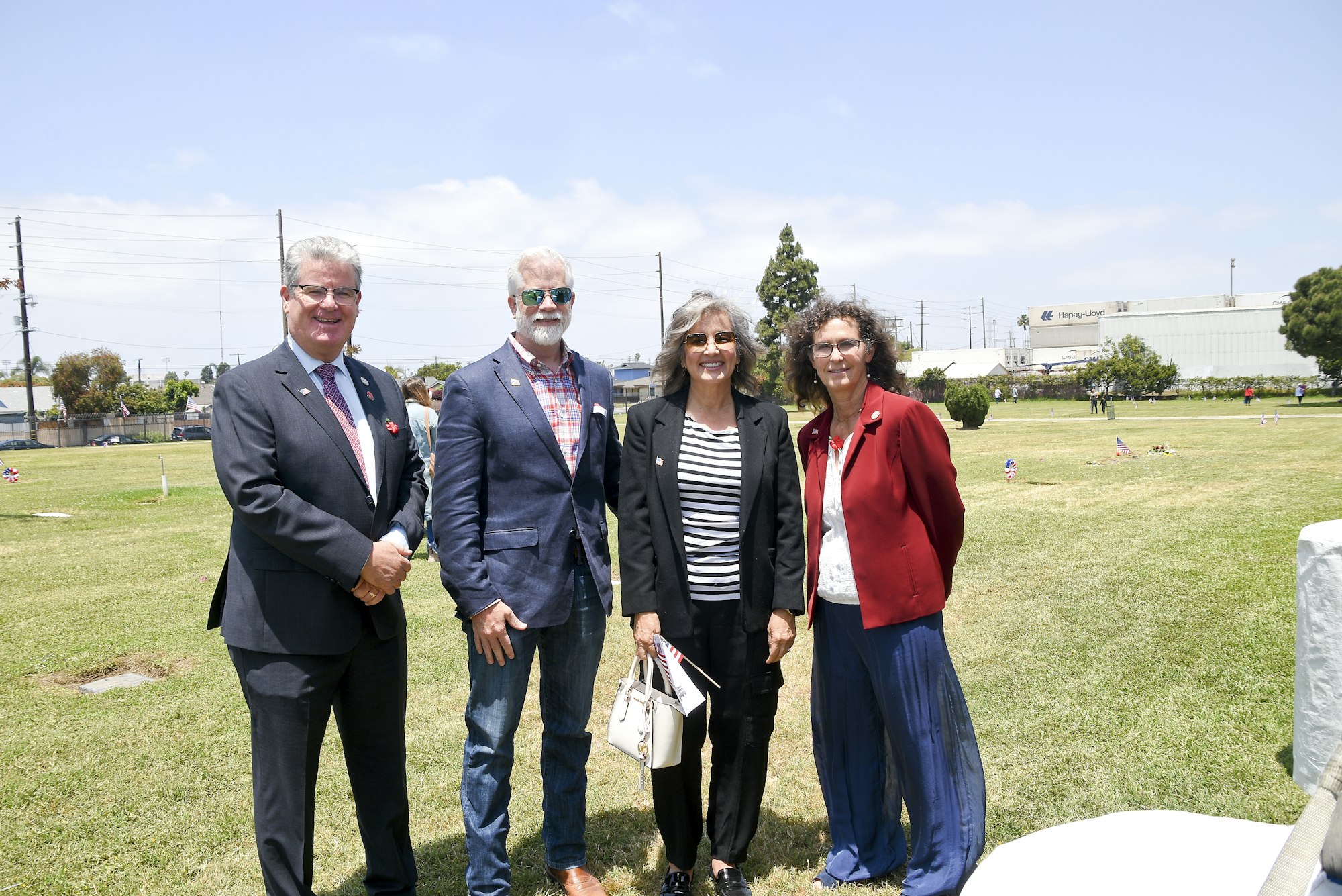 Four adults standing on grass, graves behind, industrial background, sunny day.