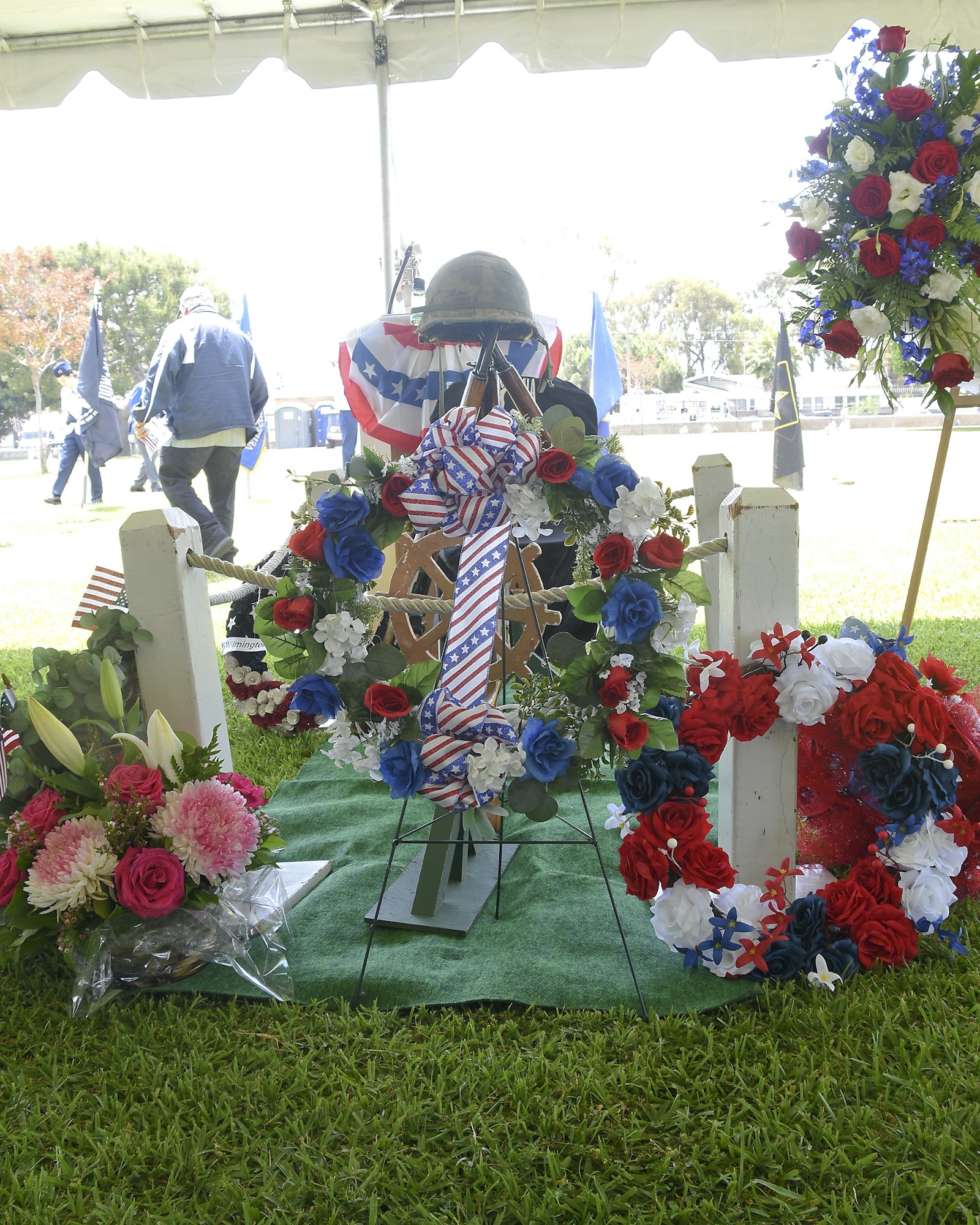 A commemorative display with a helmet, American flag, flowers, and crosses, likely at a veteran memorial event.