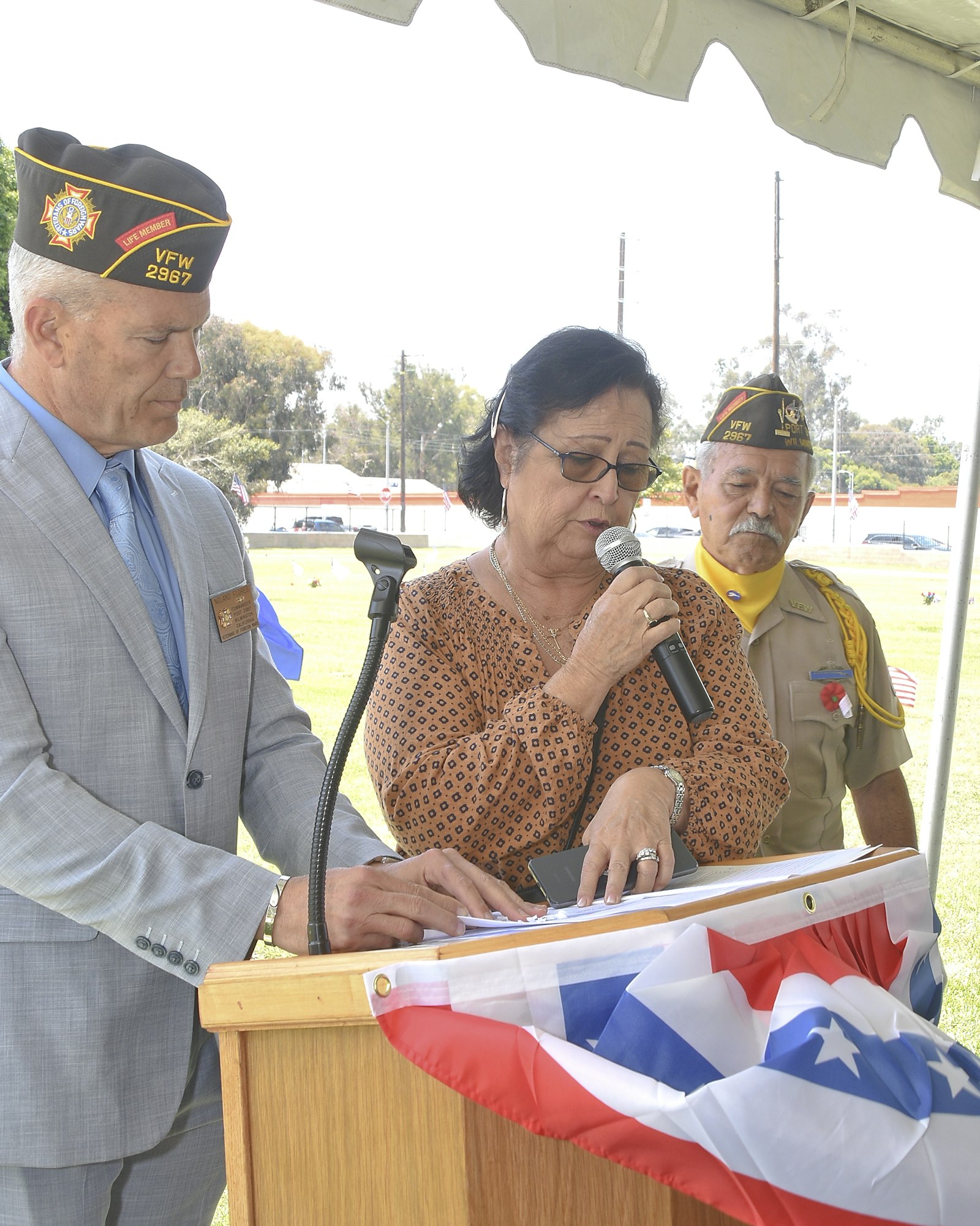 A woman speaks at a podium with a microphone, flanked by two men in VFW caps, with a folded American flag in the foreground.