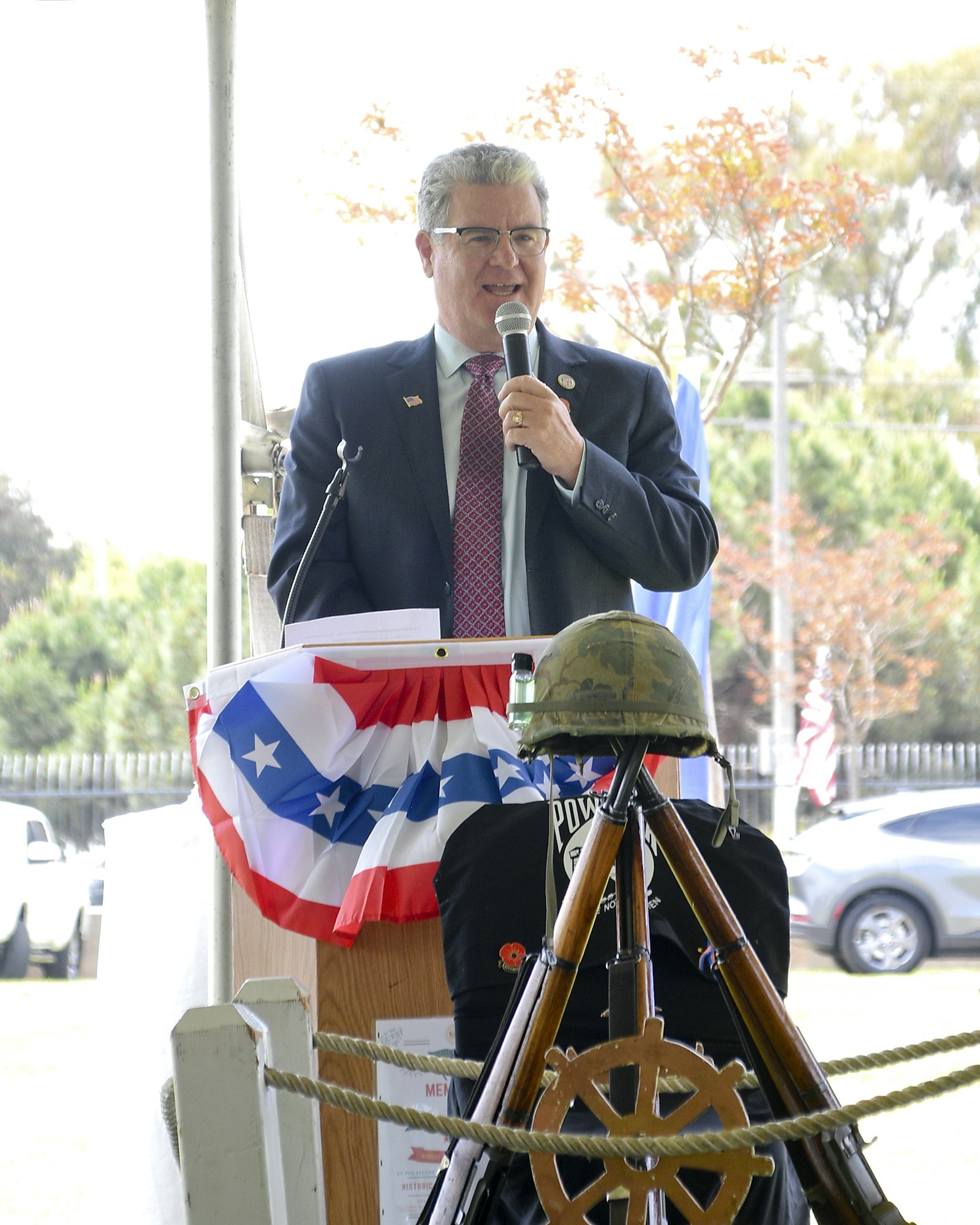 Man speaking into a microphone at a podium adorned with patriotic decor and a military helmet.