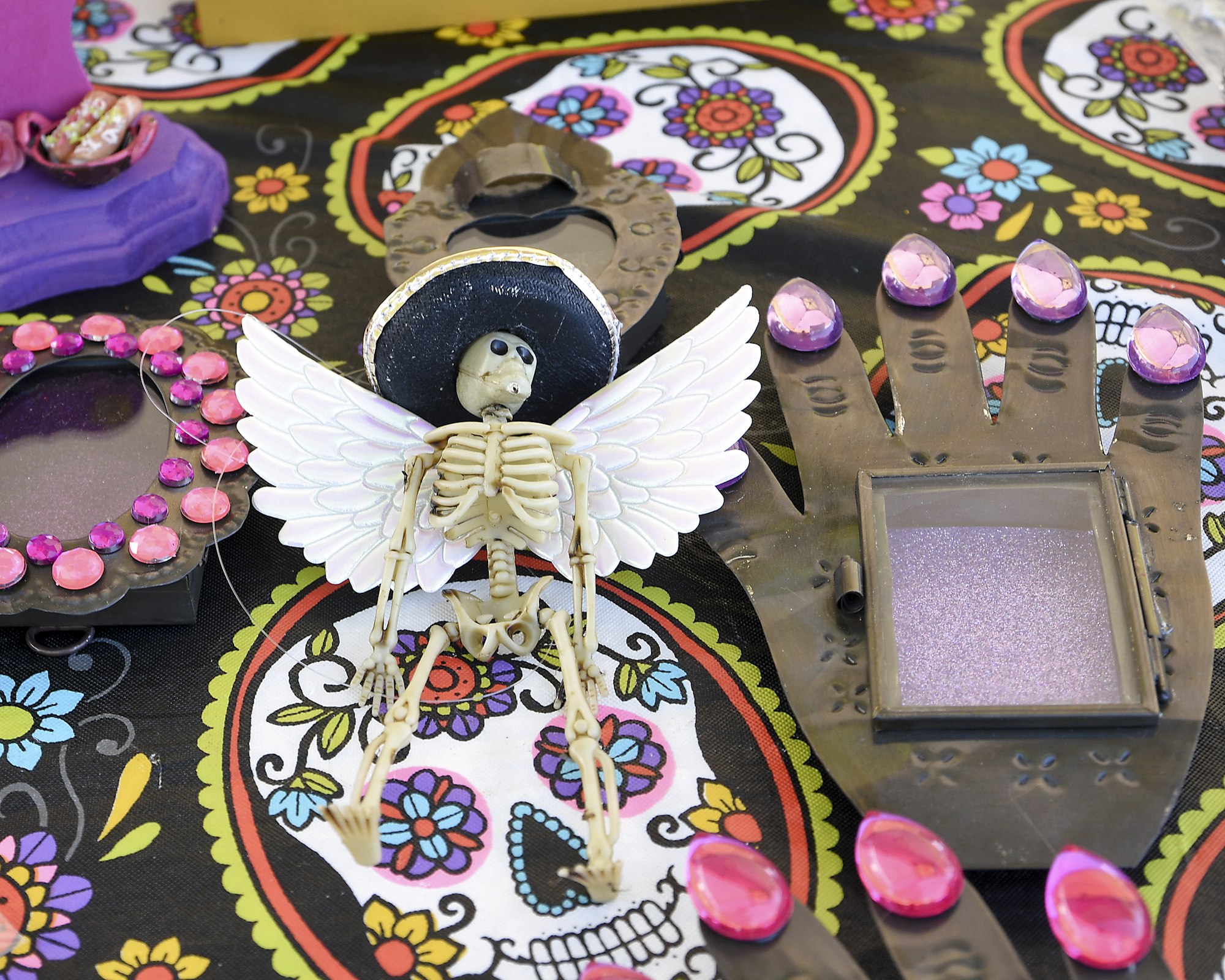 Colorful Day of the Dead-themed table display featuring handcrafted decorations. At the center is a small skeleton figure with white wings and a black hat. Surrounding it are various painted objects, including a purple heart-shaped piece and a metallic hand with pink-tipped fingers. The tablecloth is decorated with vibrant sugar skull patterns and floral designs, creating a festive and artistic atmosphere.