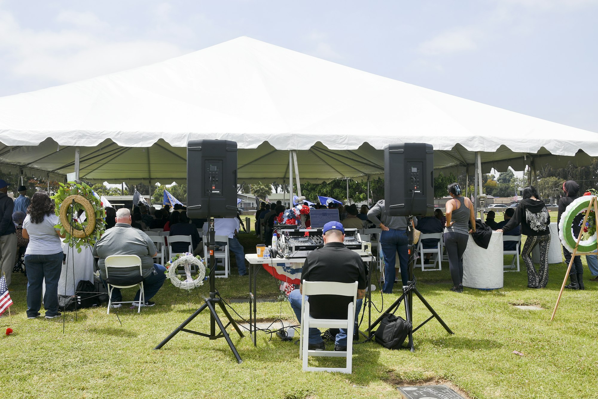 An outdoor gathering under a large tent with people seated and standing, speakers in the foreground, and wreaths displayed.