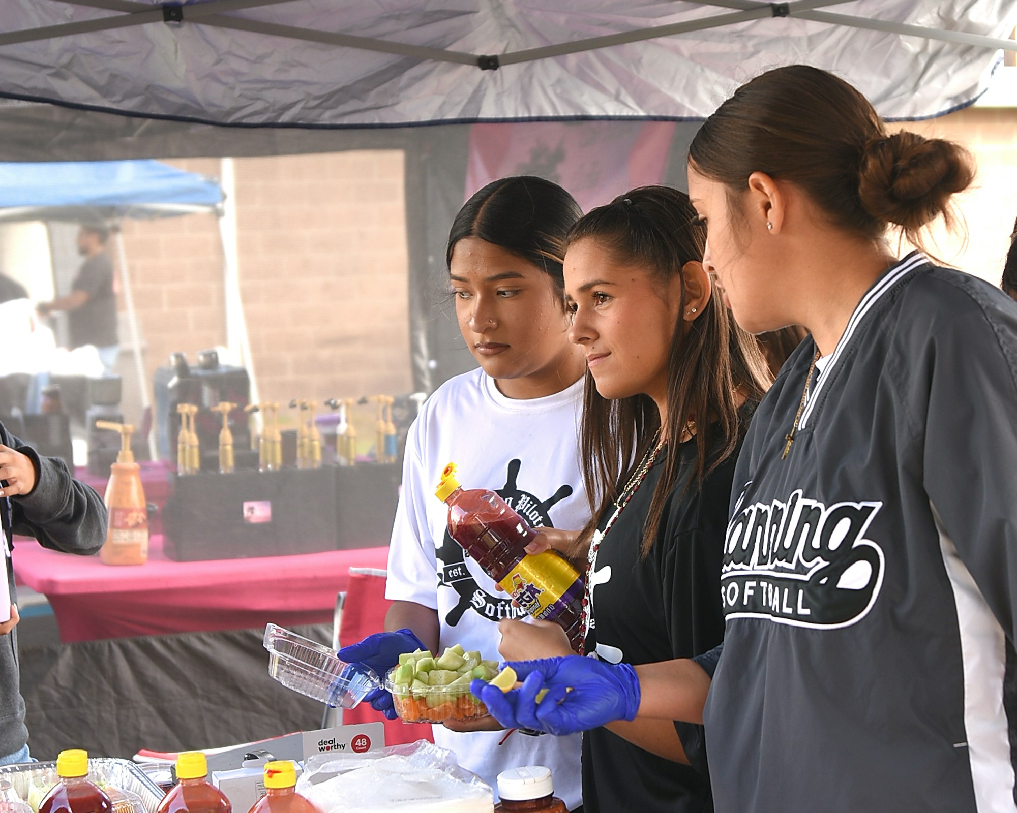 Three individuals at a food stand with drinks and condiments, under a canopy tent.