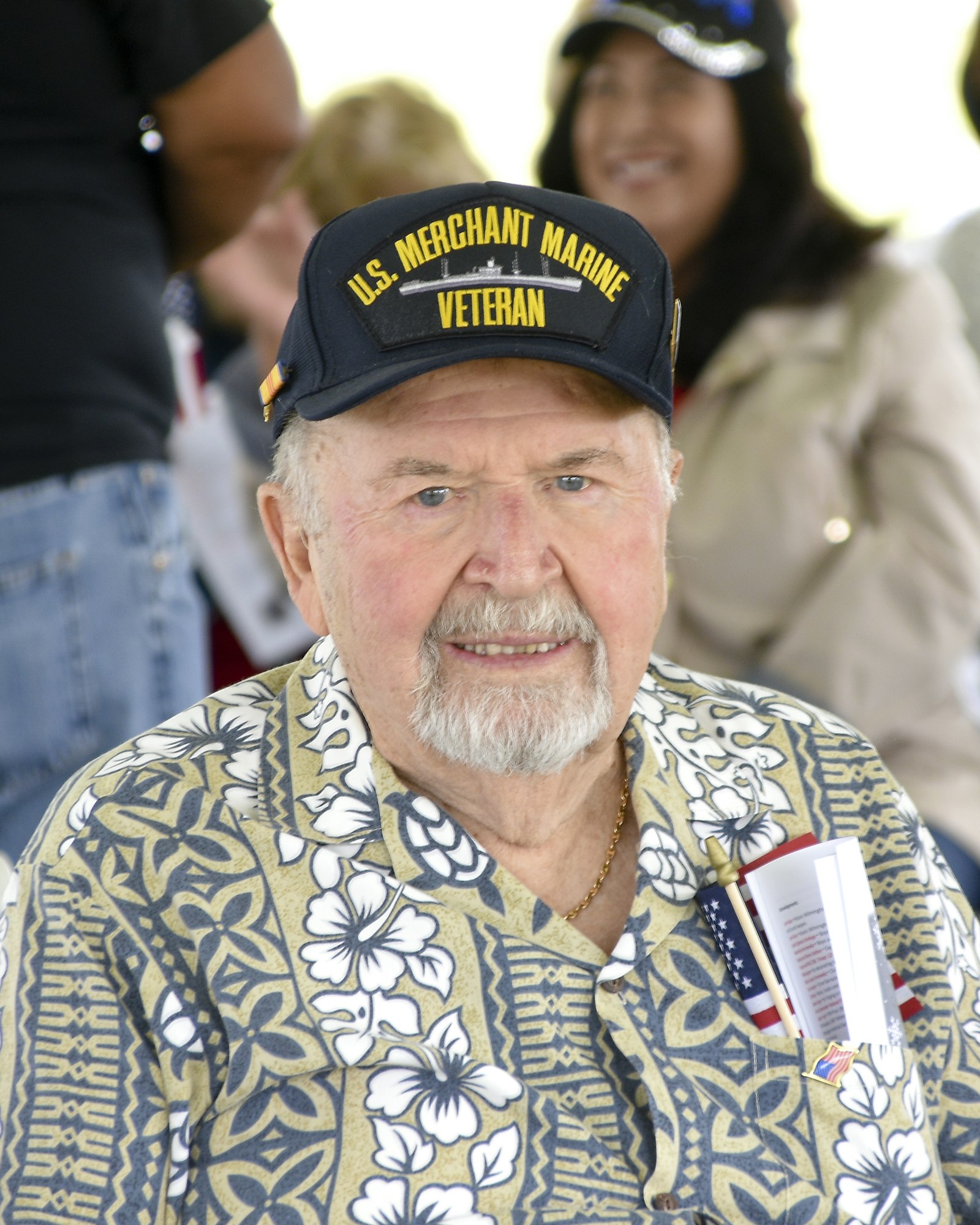 An elderly man in a floral shirt wearing a "U.S. Merchant Marine Veteran" cap with an American flag pin and program.
