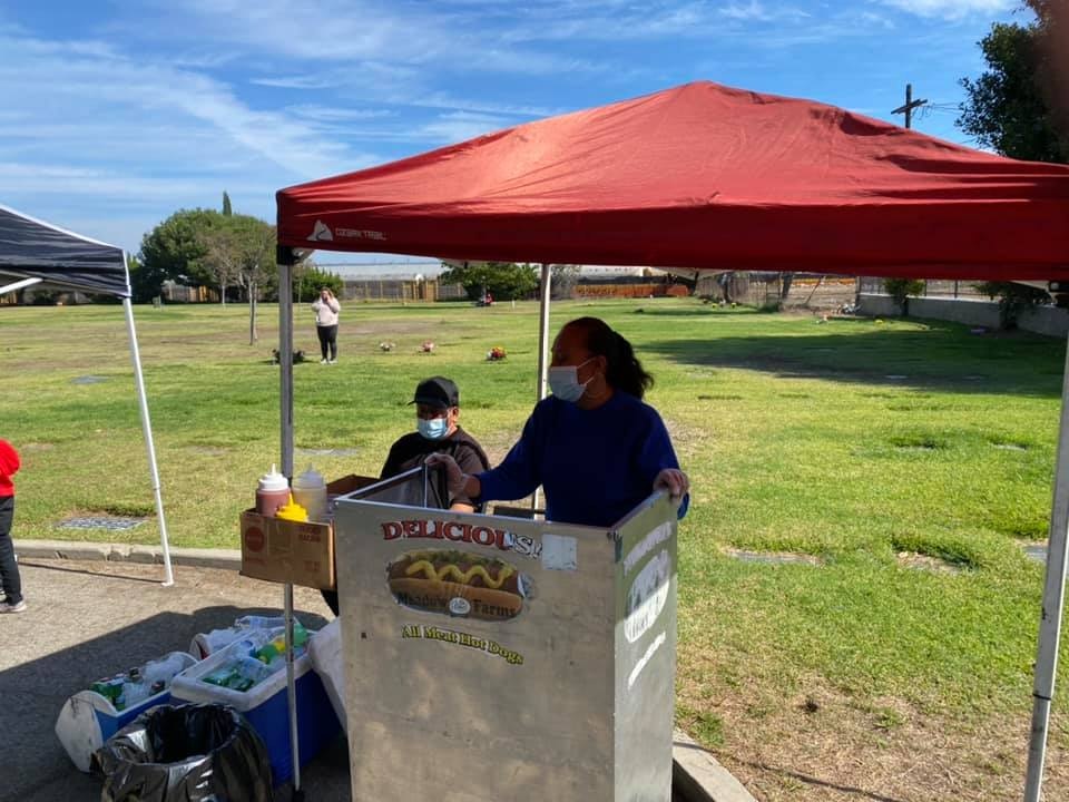 A food vendor under a red tent serves hot dogs in a park, with people in the background enjoying the outdoors.
