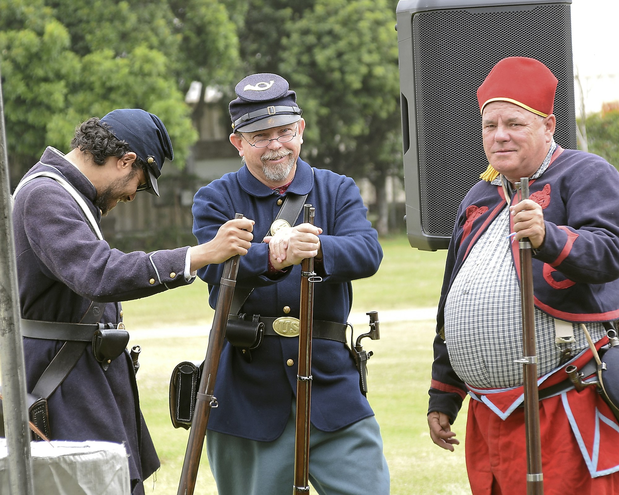 Three people in historical costume, two with rifles, and one wearing a fez-like hat, appear to be at a reenactment.