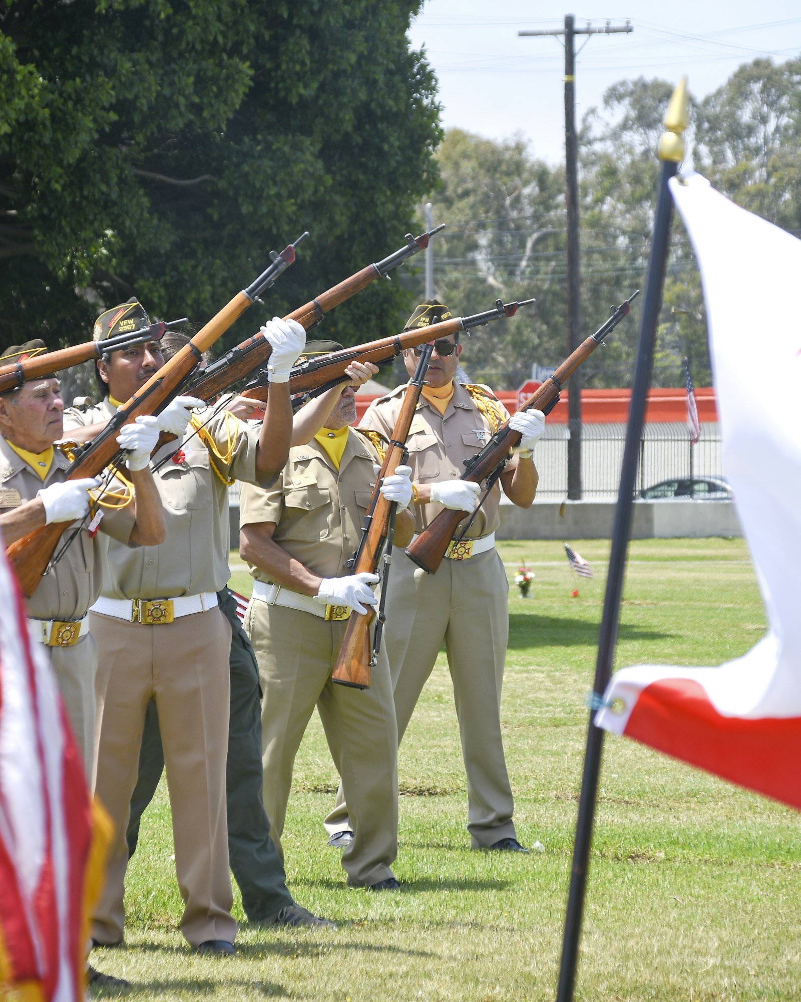 Uniformed individuals performing a rifle salute near flags, possibly at a ceremonial event.