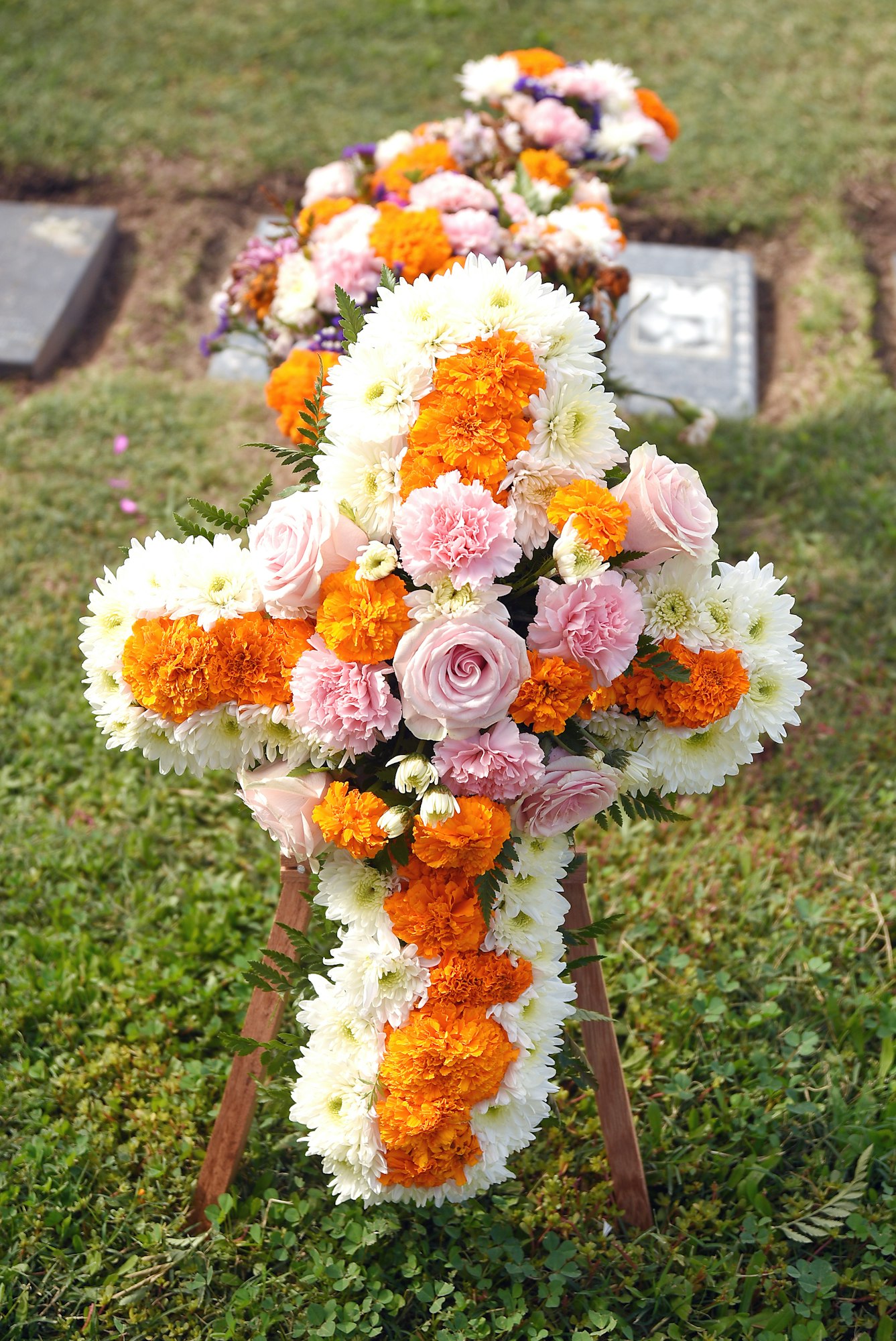 A floral cross arrangement with white, orange, and pink blooms standing in a grassy field, possibly a cemetery.