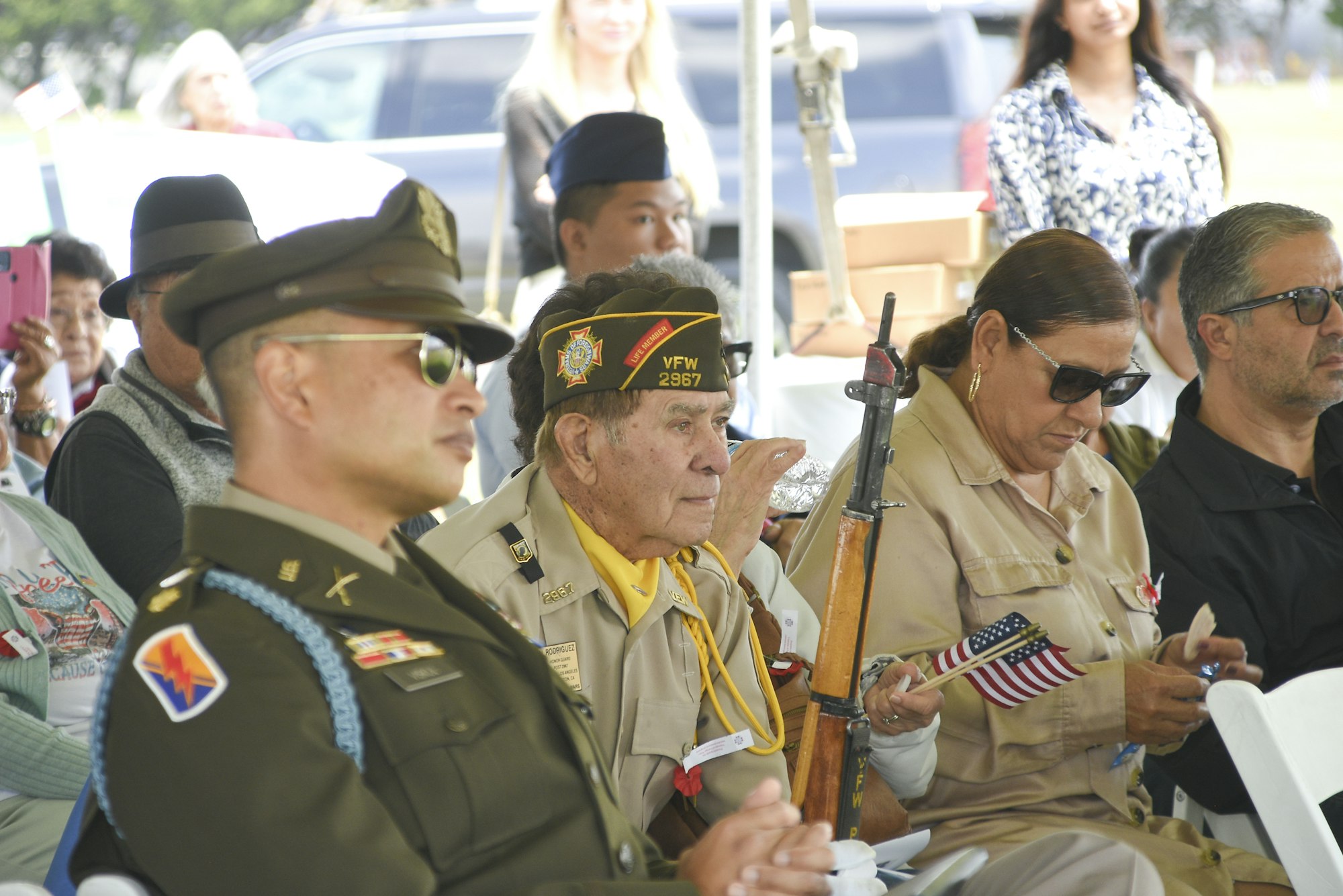 Military personnel and civilians at an event, one holding a small American flag, indicating a patriotic or commemorative gathering.