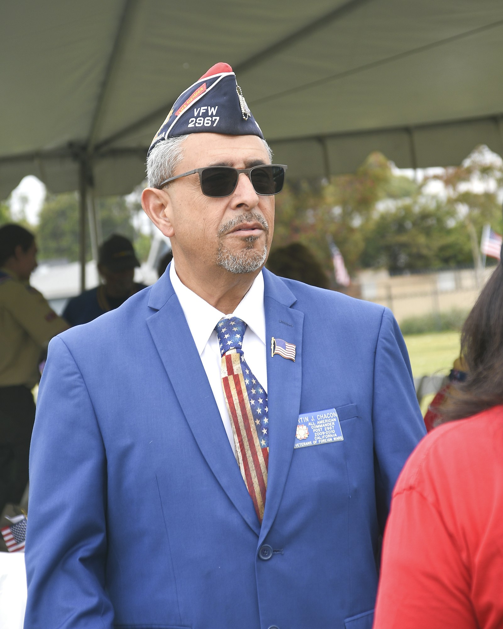 Man in blue suit, VFW cap, sunglasses, patriotic tie, at an outdoor event.