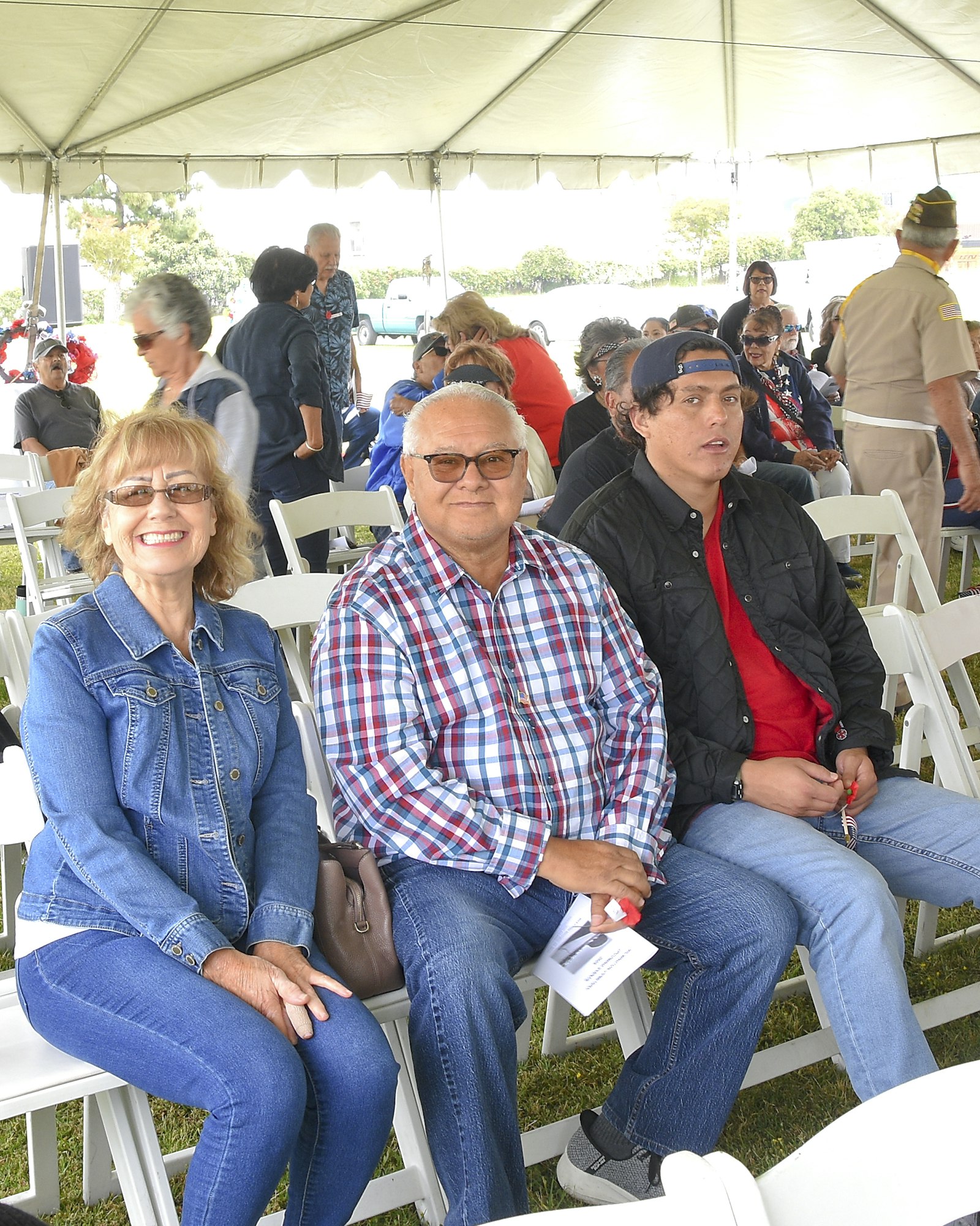 Three people sitting in chairs under a tent at an outdoor event.