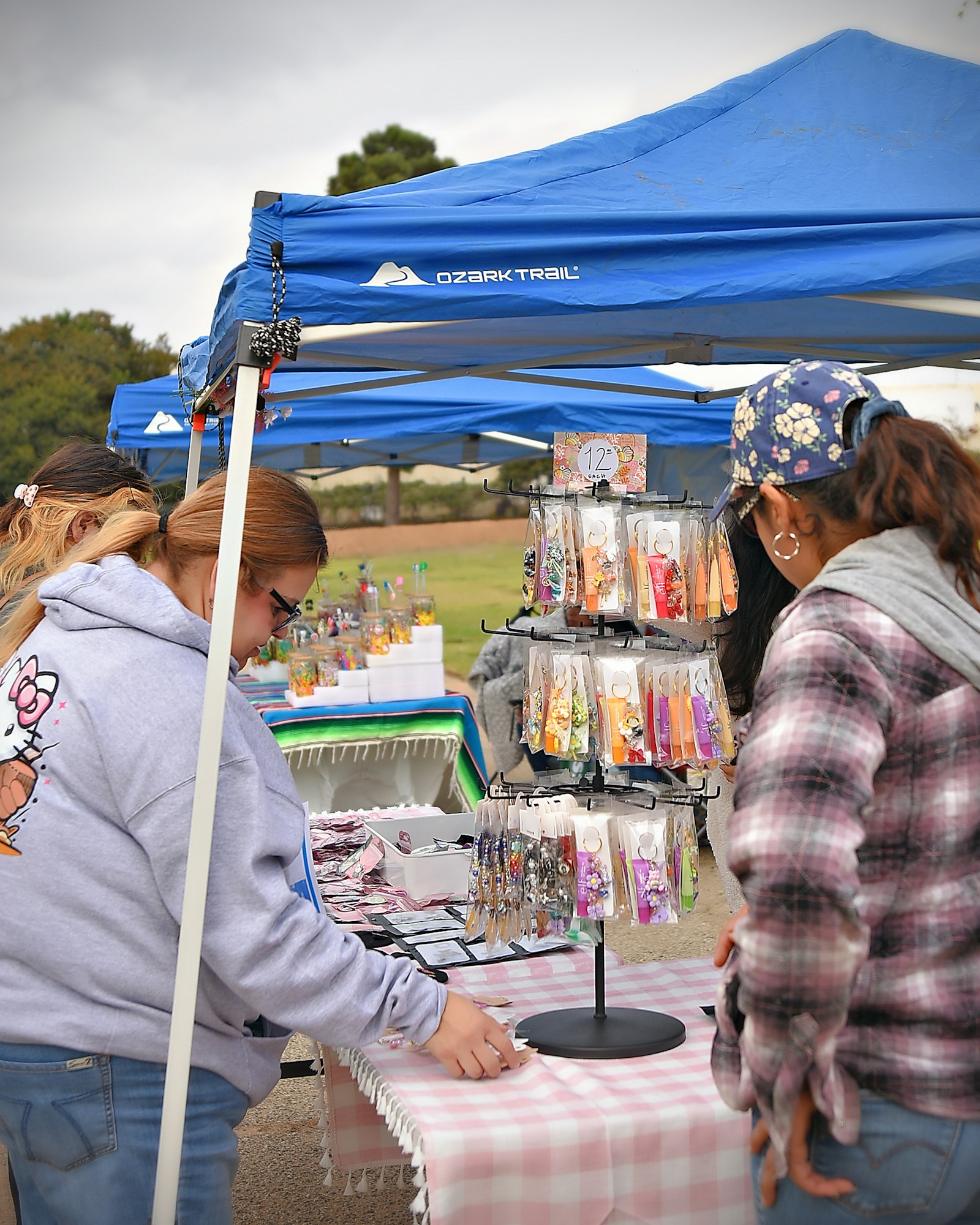 An outdoor vendor stall with people browsing earrings under a blue canopy.