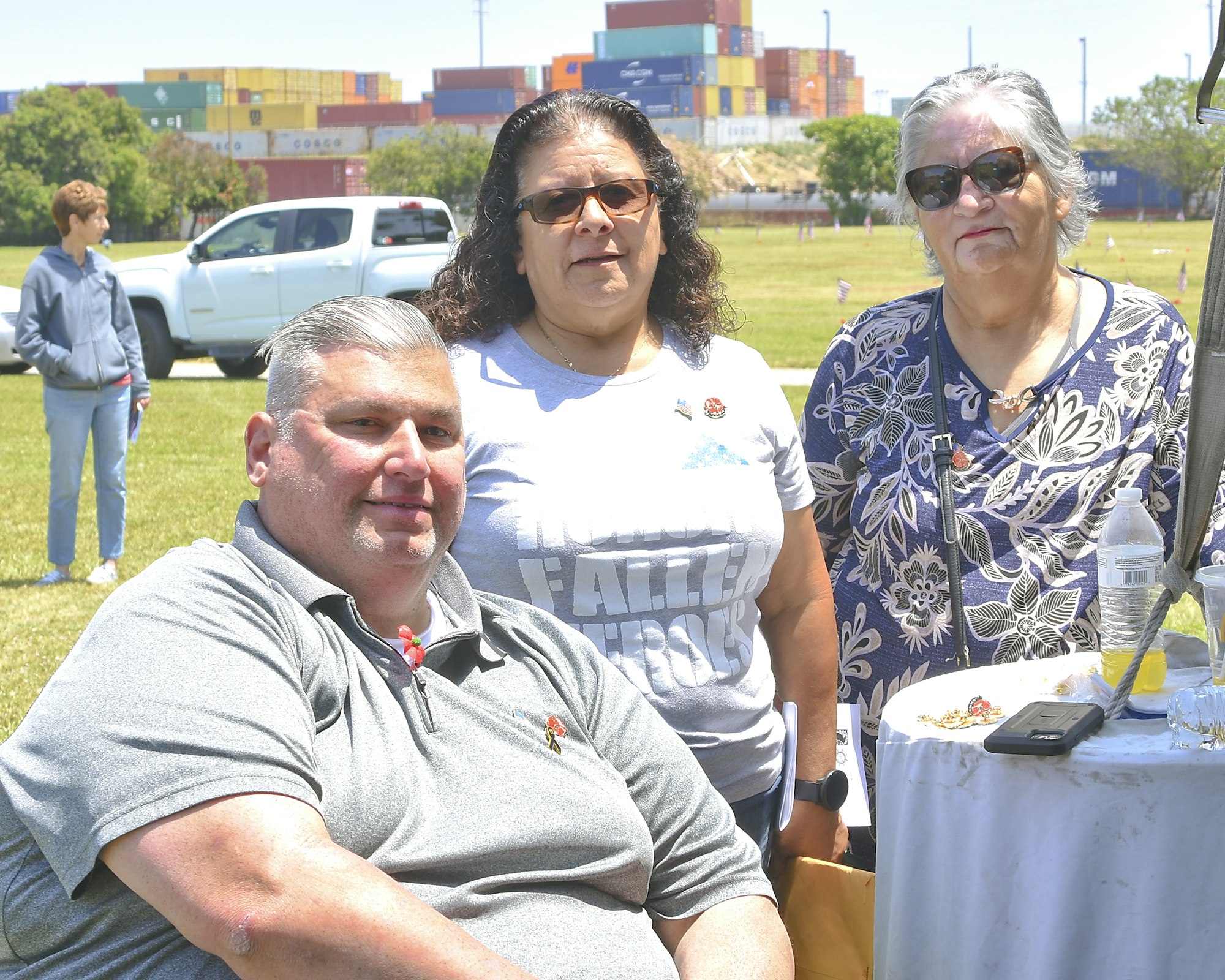 Three people posing at a table outdoors with shipping containers in the background.